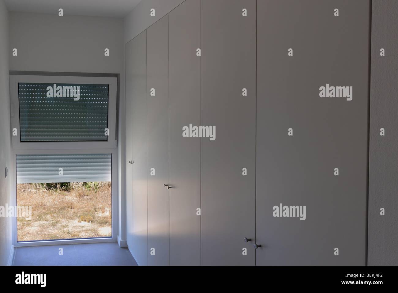 Une chambre propre et contemporaine dispose d'une armoire blanche du sol au plafond avec des poignées subtiles et une grande fenêtre révélant un SC extérieur ensoleillé et aride Banque D'Images