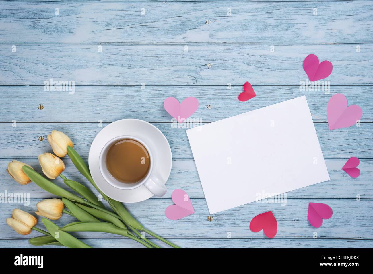 Une scène chaleureuse avec une tasse de café, des tulipes jaunes et une carte vierge entourée de coeurs roses sur un fond de bois, parfait pour la fête des mères Banque D'Images