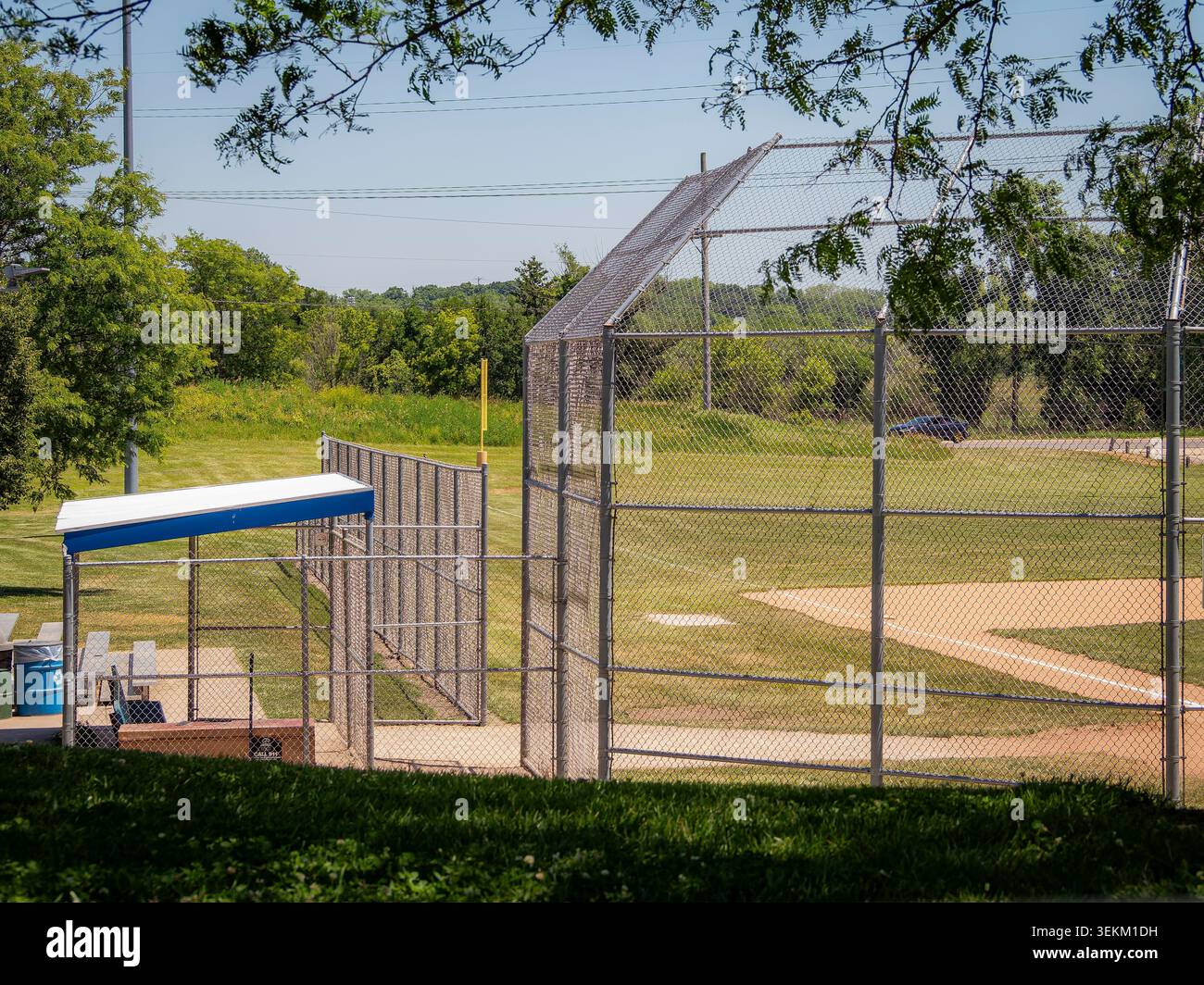 Cage vide de backstop de terrain de baseball et dugout dans un parc communautaire par une journée ensoleillée brillante avec des arbres verts et un fond de ciel bleu Banque D'Images