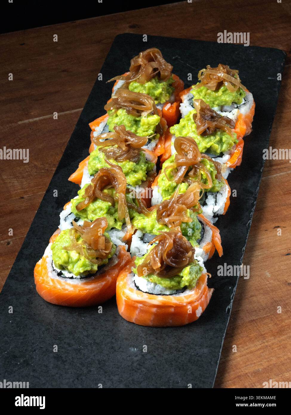 Rouleaux de saumon frais recouverts de guacamole et d'oignons caramélisés. Servi sur un élégant plateau en pierre noire. La table en bois chaude améliore la présentation rustique Banque D'Images