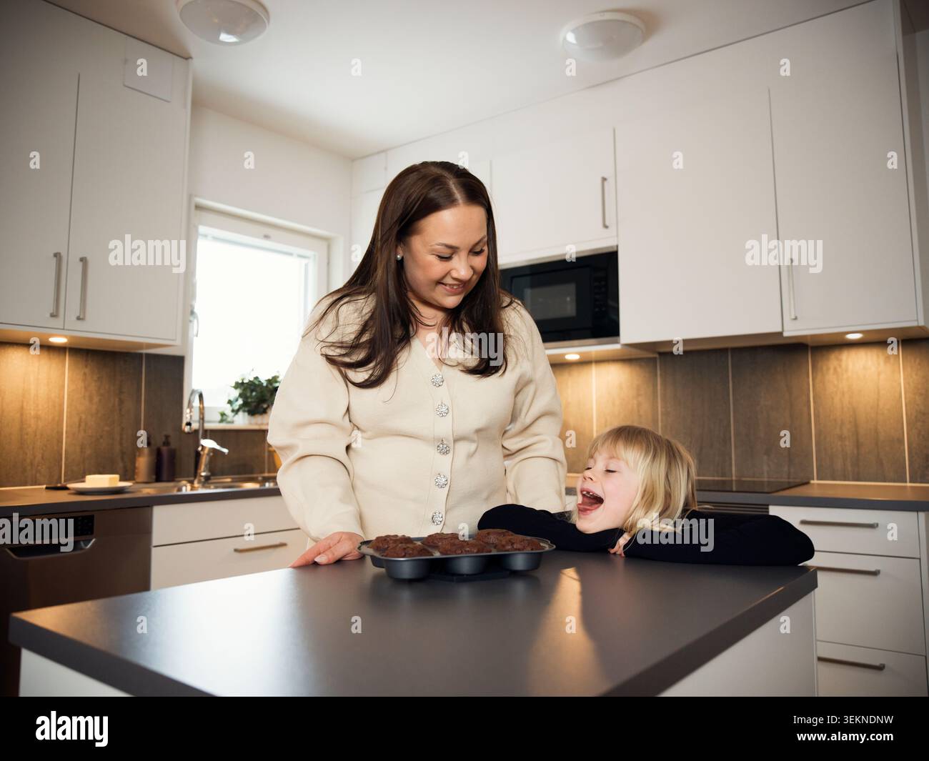 Fille ludique debout avec mère près de la boîte à muffins gardée sur la table à la maison Banque D'Images