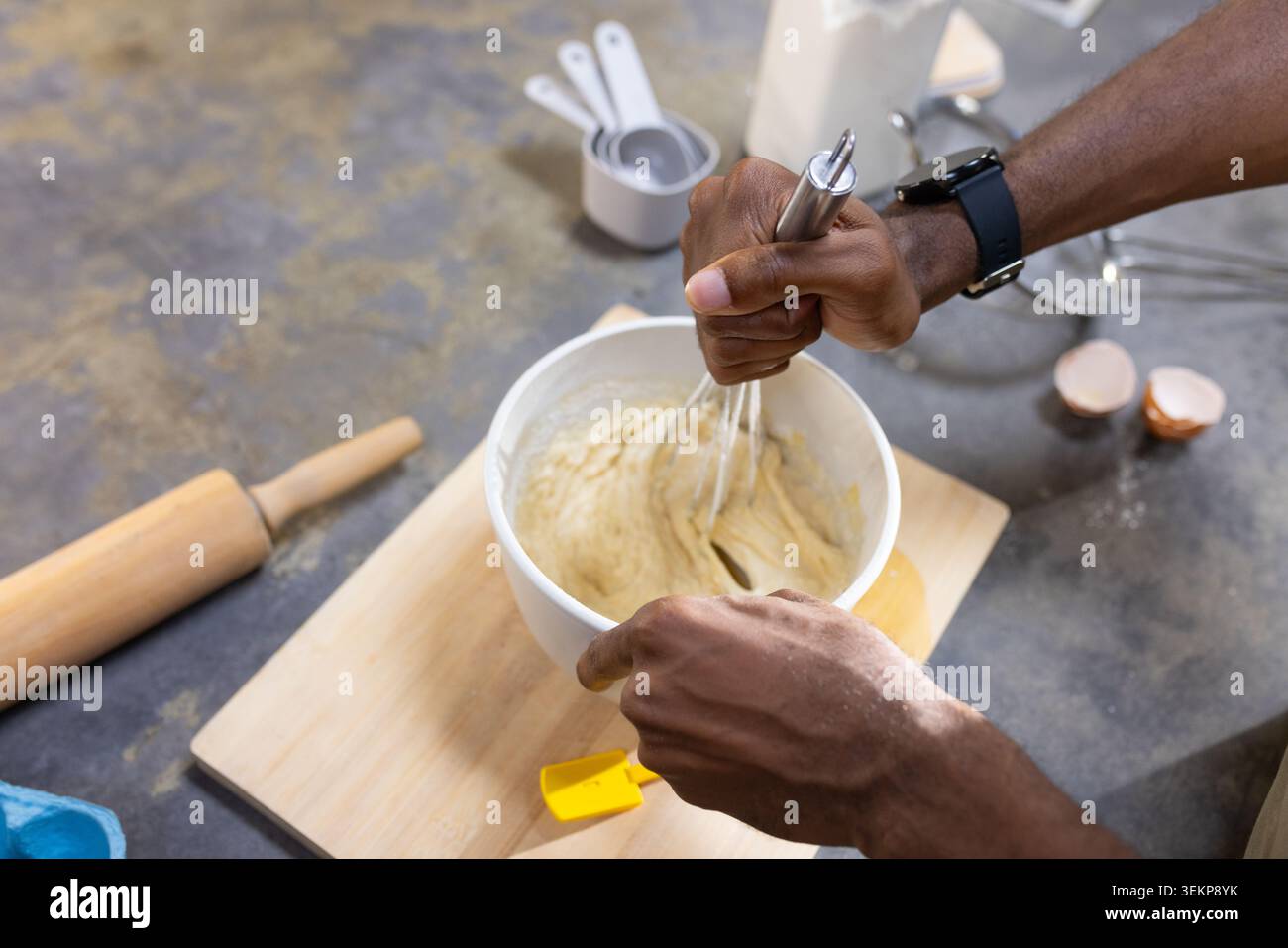 Homme fouettant la pâte avec un fouet en métal dans le bol à mélanger sur le comptoir de la cuisine, tenant la spatule jaune Banque D'Images
