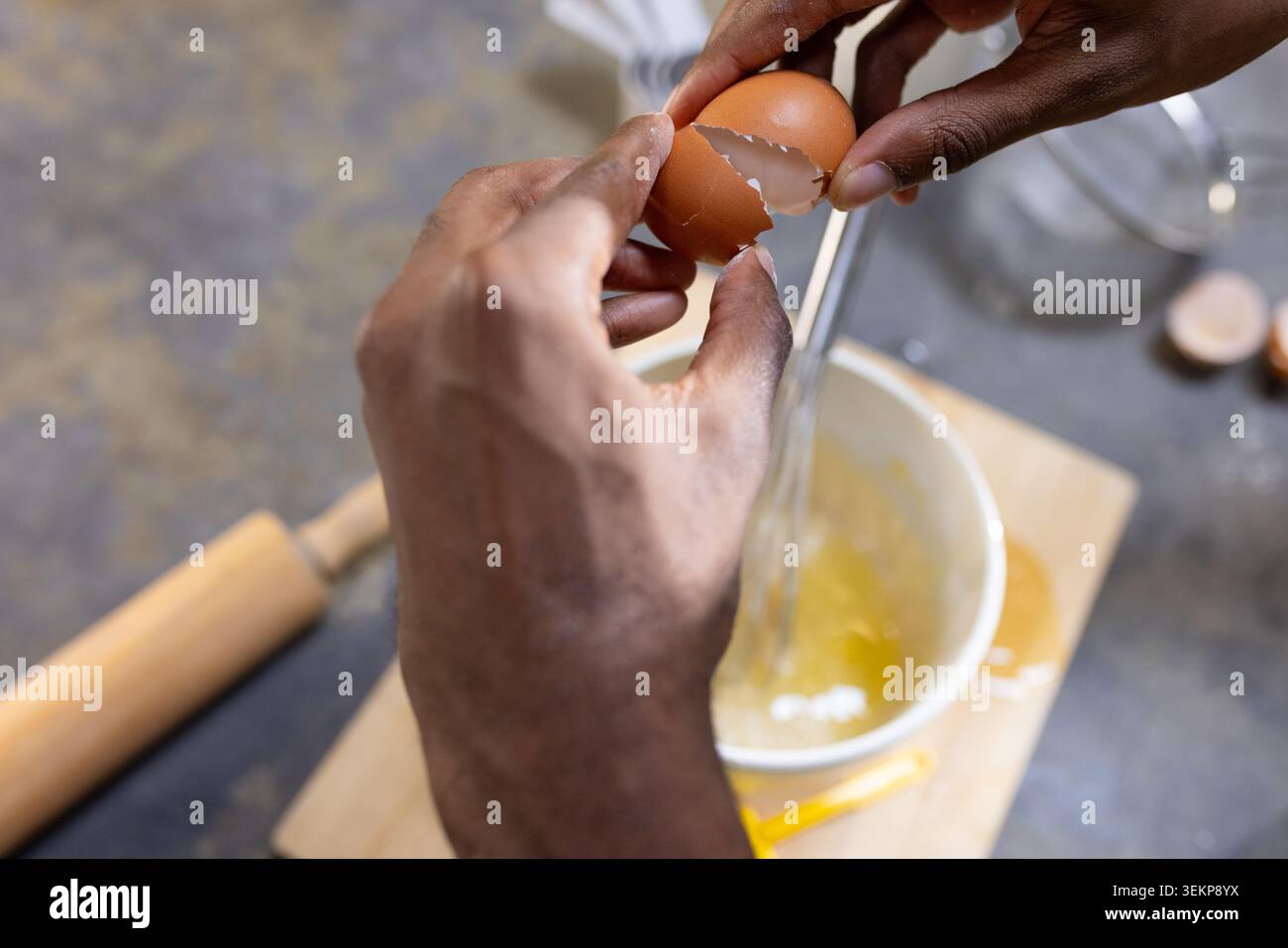 Mâle africain adulte cassant l'oeuf brun dans un bol à mélanger en céramique avec un fouet en métal sur la planche à découper Banque D'Images