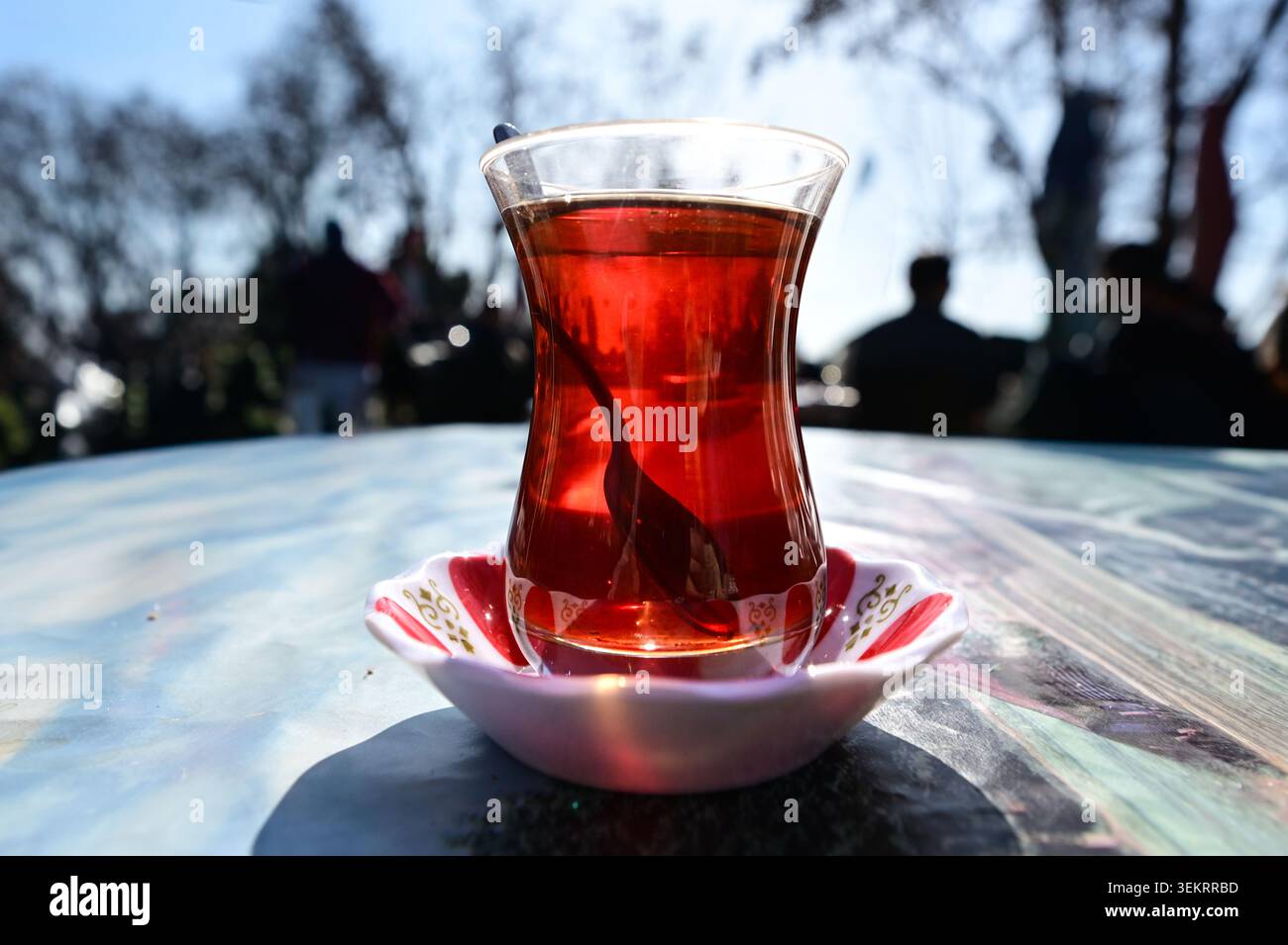 Thé turc rétro-éclairé dans un verre de tulipe sur une soucoupe, table de café en plein air avec une faible profondeur de champ et des reflets rouges chauds. Banque D'Images