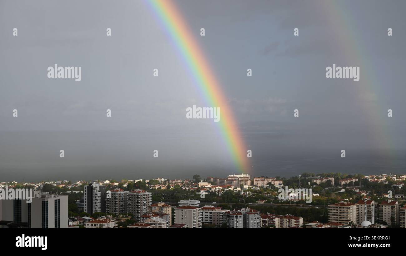 Arc-en-ciel vif et arc-en-ciel secondaire faible voûtant au-dessus de l'horizon d'une ville côtière, de la mer et du ciel nuageux. Banque D'Images