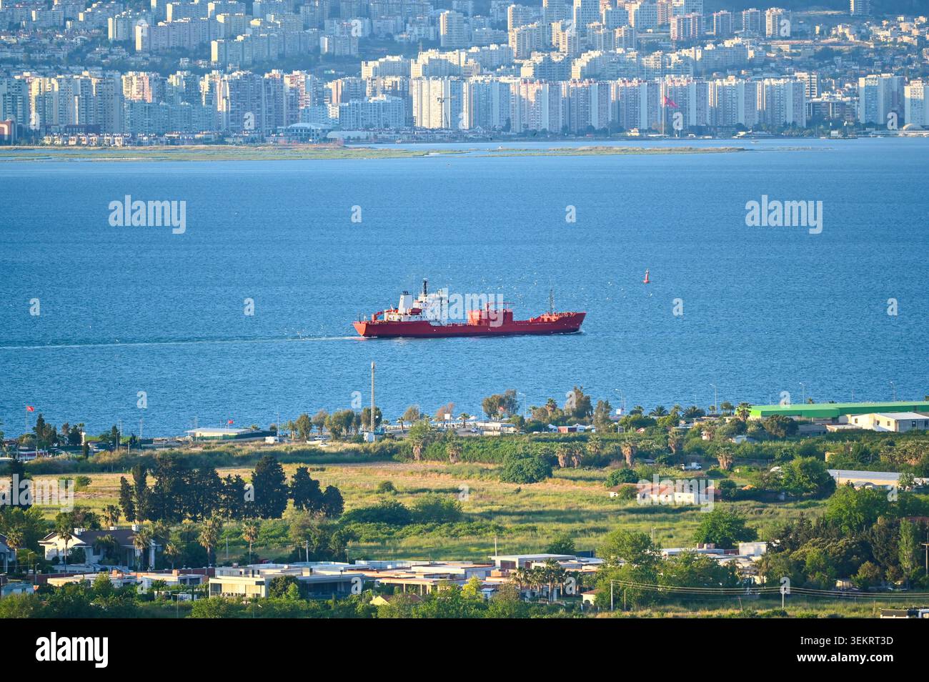 Navire de charge rouge naviguant dans la mer Égée avec le skyline lointain de la ville d'Izmir et des tours résidentielles sous une lumière diurne éclatante. Banque D'Images