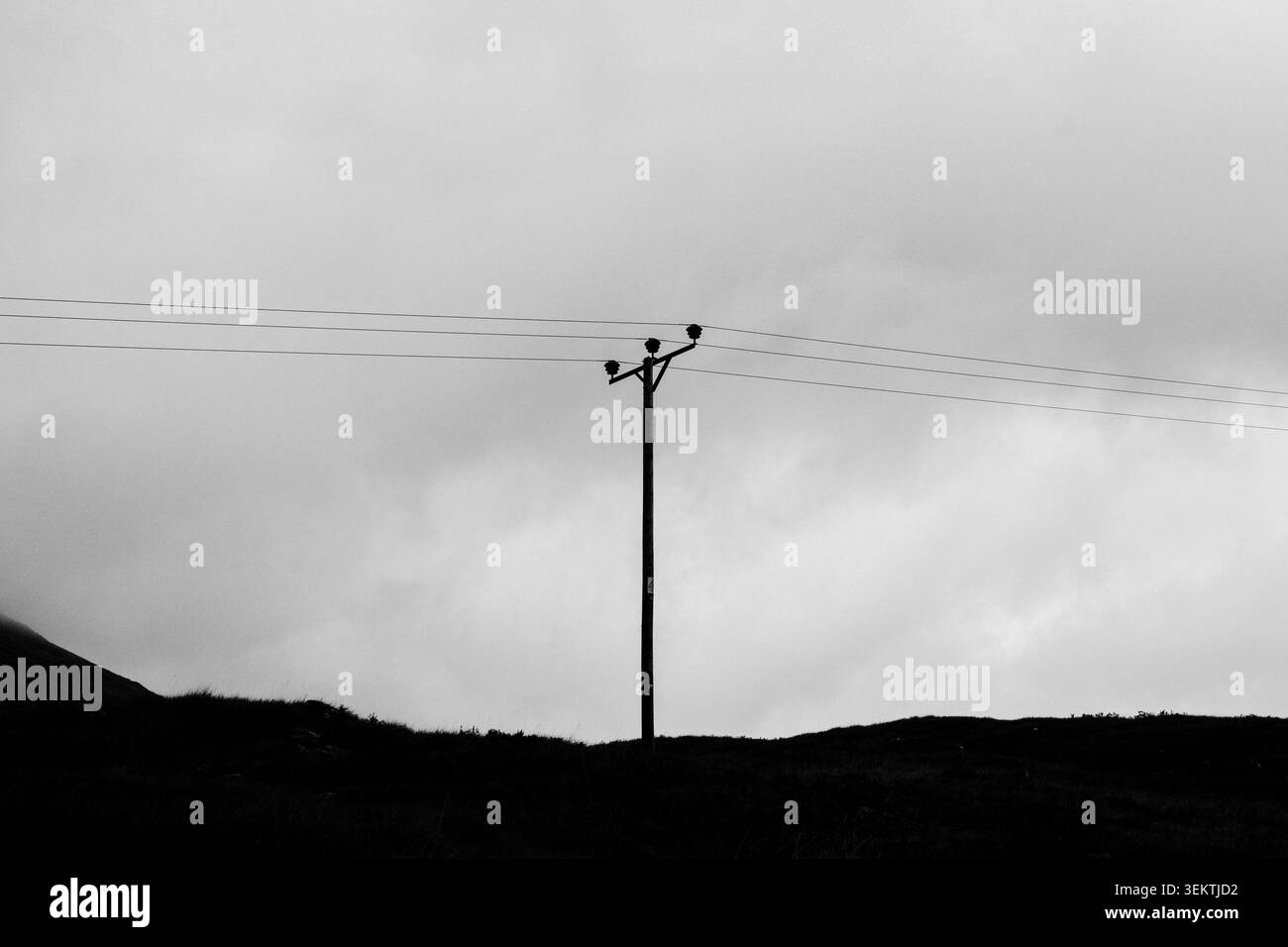 Paysage monochrome graphique avec un pylône électrique unique et des fils aériens contre un ciel nuageux couvert, composition minimaliste spectaculaire. Banque D'Images