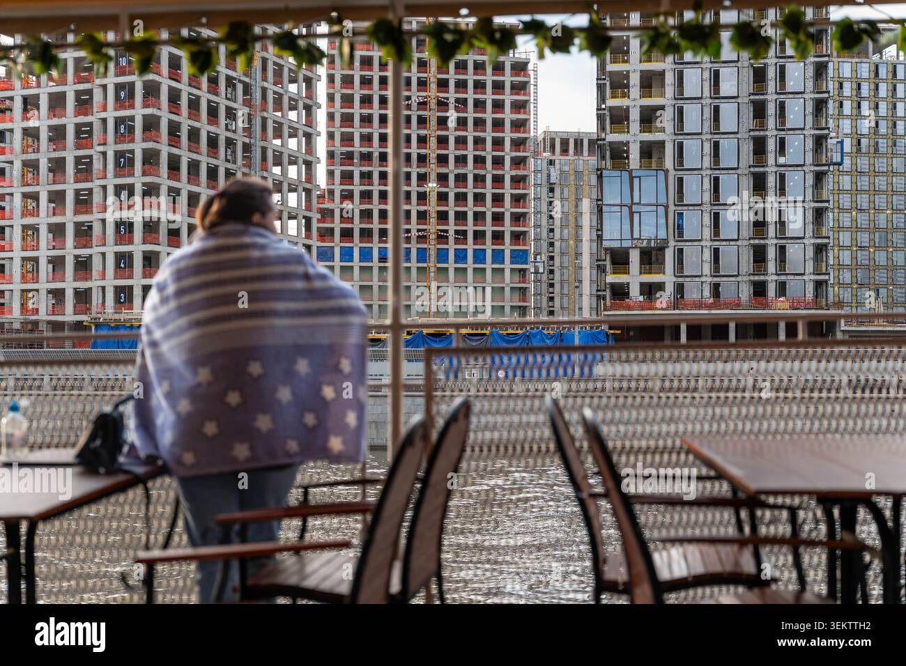 Personne enveloppée dans une couverture sur le balcon surplombant la rivière et le chantier de construction, chaises et tables en métal vides, Cold Morning Haze, appartement urbain Banque D'Images