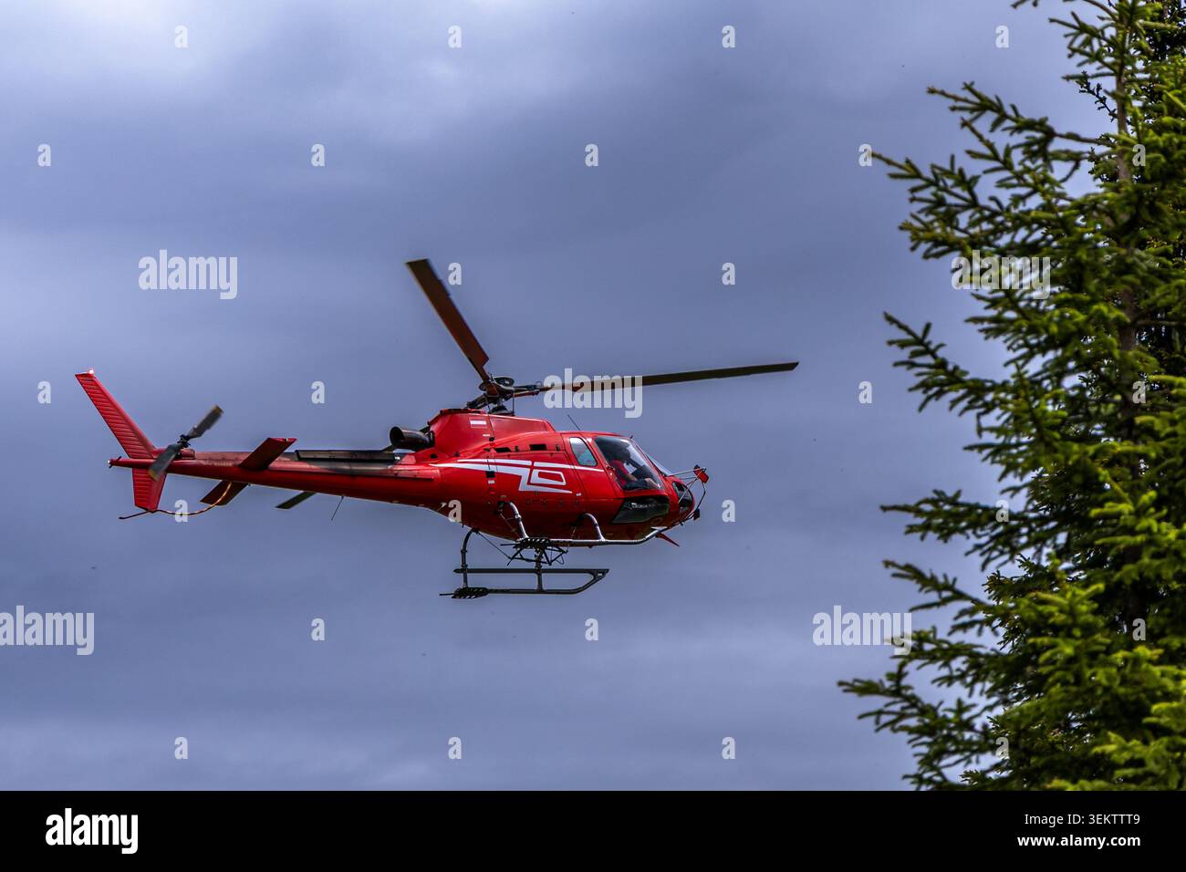 Un hélicoptère rouge éclatant survole un ciel nuageux au-dessus d'un paysage de montagne verdoyant et luxuriant. L'hélicoptère est capturé à mi-vol, montrant son powe Banque D'Images