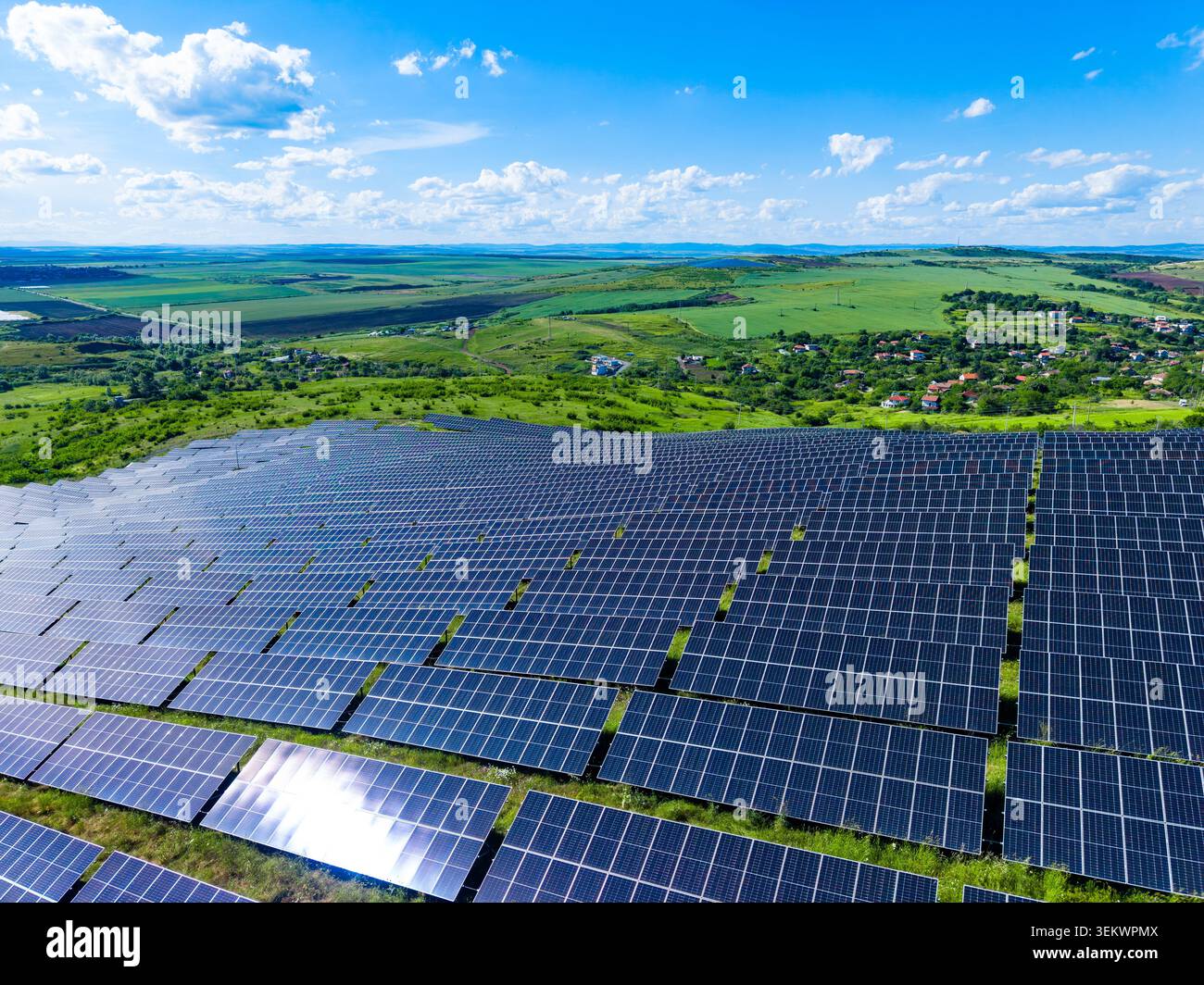 Les panneaux solaires couvrent une grande surface dans un champ. Des collines verdoyantes et quelques petites maisons sont visibles au loin. La scène est lumineuse avec un ciel bleu et blanc Banque D'Images