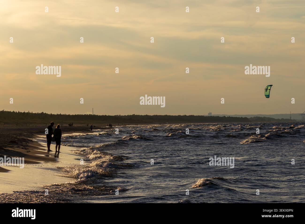 Sobiszewska Island, POLOGNE-SEPTEMBRE 02, 2024 : couple marchant le long d'une plage de sable au coucher du soleil, profitant de la mer Baltique ondulée tandis qu'un kite surfeur est coloré Banque D'Images