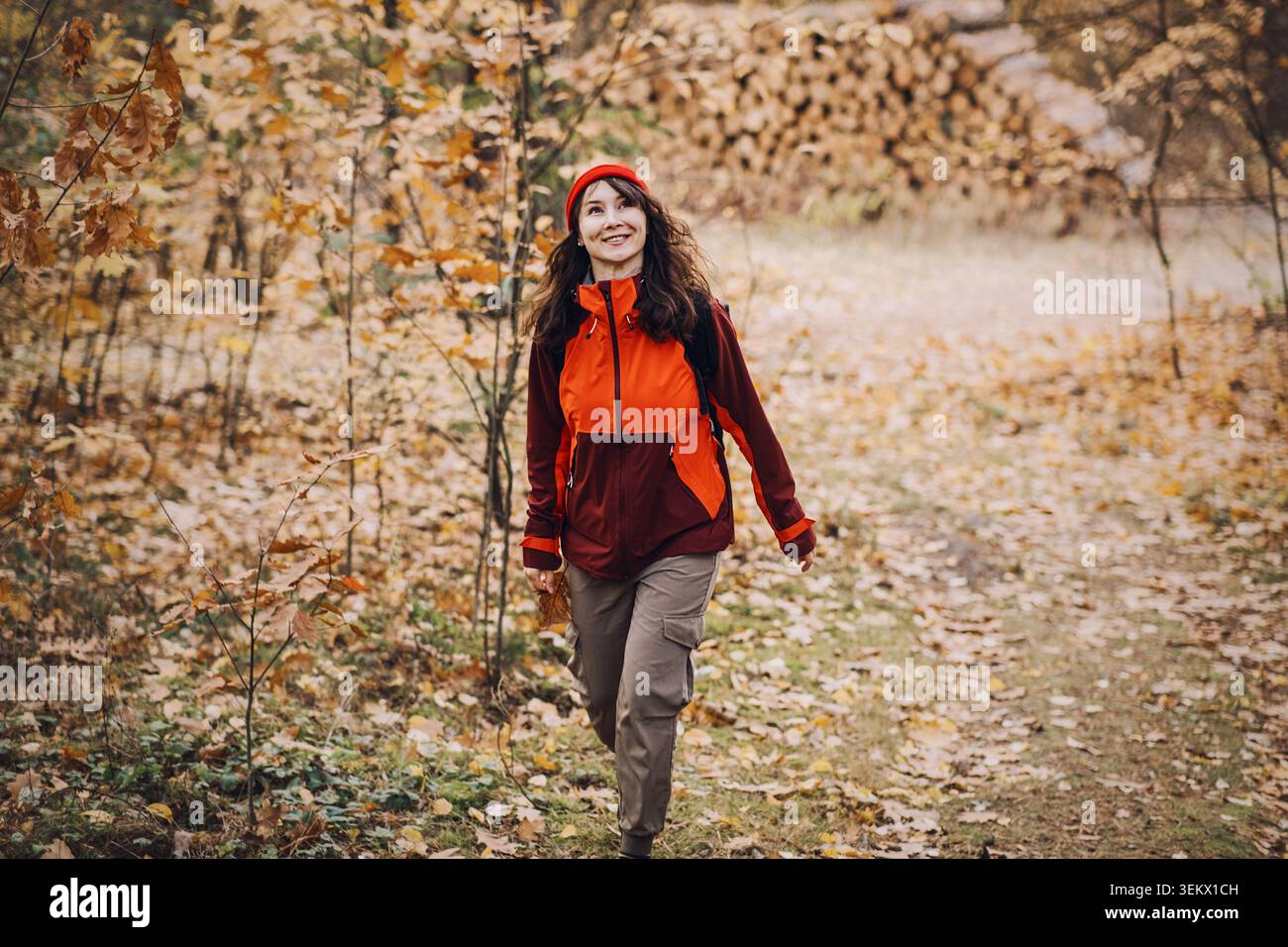 Femme souriante marche à travers la forêt d'automne devant des bûches empilées. Détox numérique dans la nature, échapper au stress urbain, vacances sans technologie. Banque D'Images