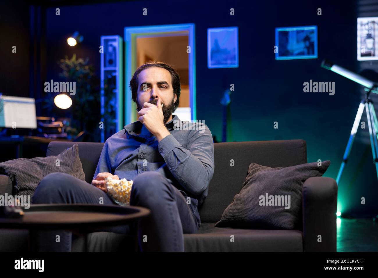 Homme paresseux appréciant le temps libre à la maison regarder la télévision, grignoter sur pop-corn. Canapé pomme de terre dans un appartement éclairé au néon, bouche farce avec collation salée, diverti par la télévision Banque D'Images