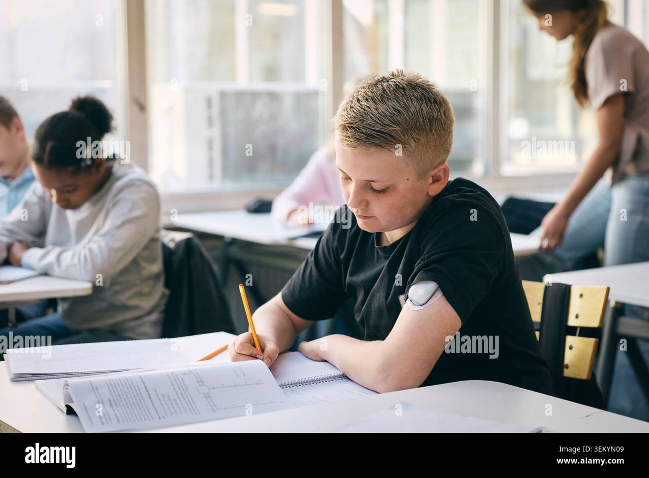 Étudiant masculin portant un moniteur de glucose sur le bras écrivant dans le livre pendant ses études à l'école Banque D'Images