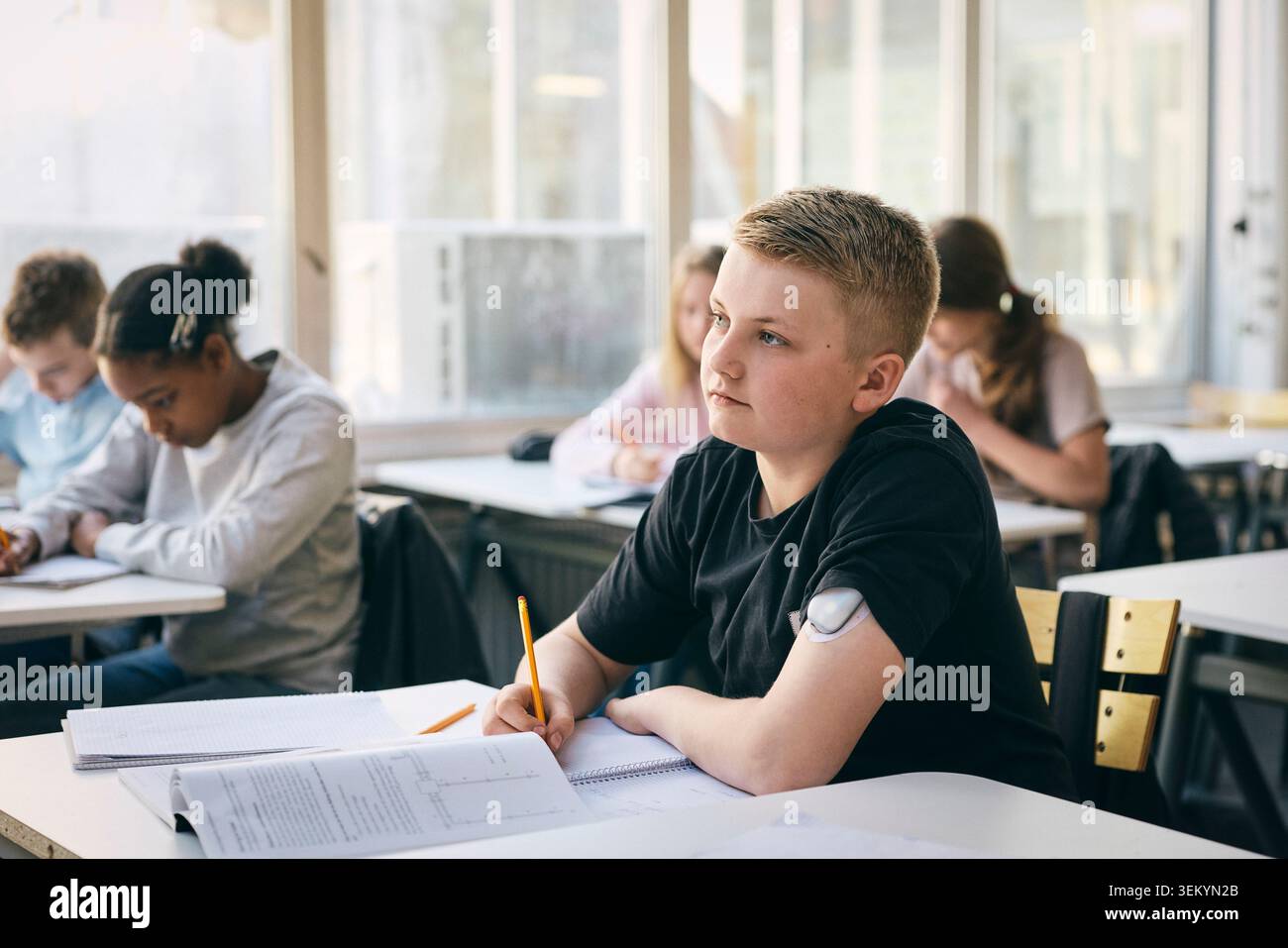 Étudiant masculin réfléchi portant un moniteur de glucose sur le bras assis au bureau pendant ses études à l'école Banque D'Images