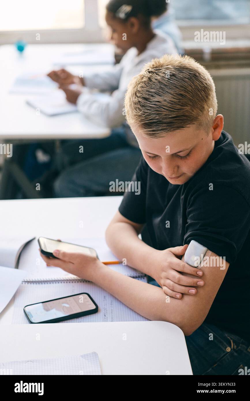 Étudiant junior de niveau élevé vérifiant le taux de sucre dans le sang à l'aide d'un glucomètre tout en étant assis au bureau avec des livres dans la salle de classe Banque D'Images