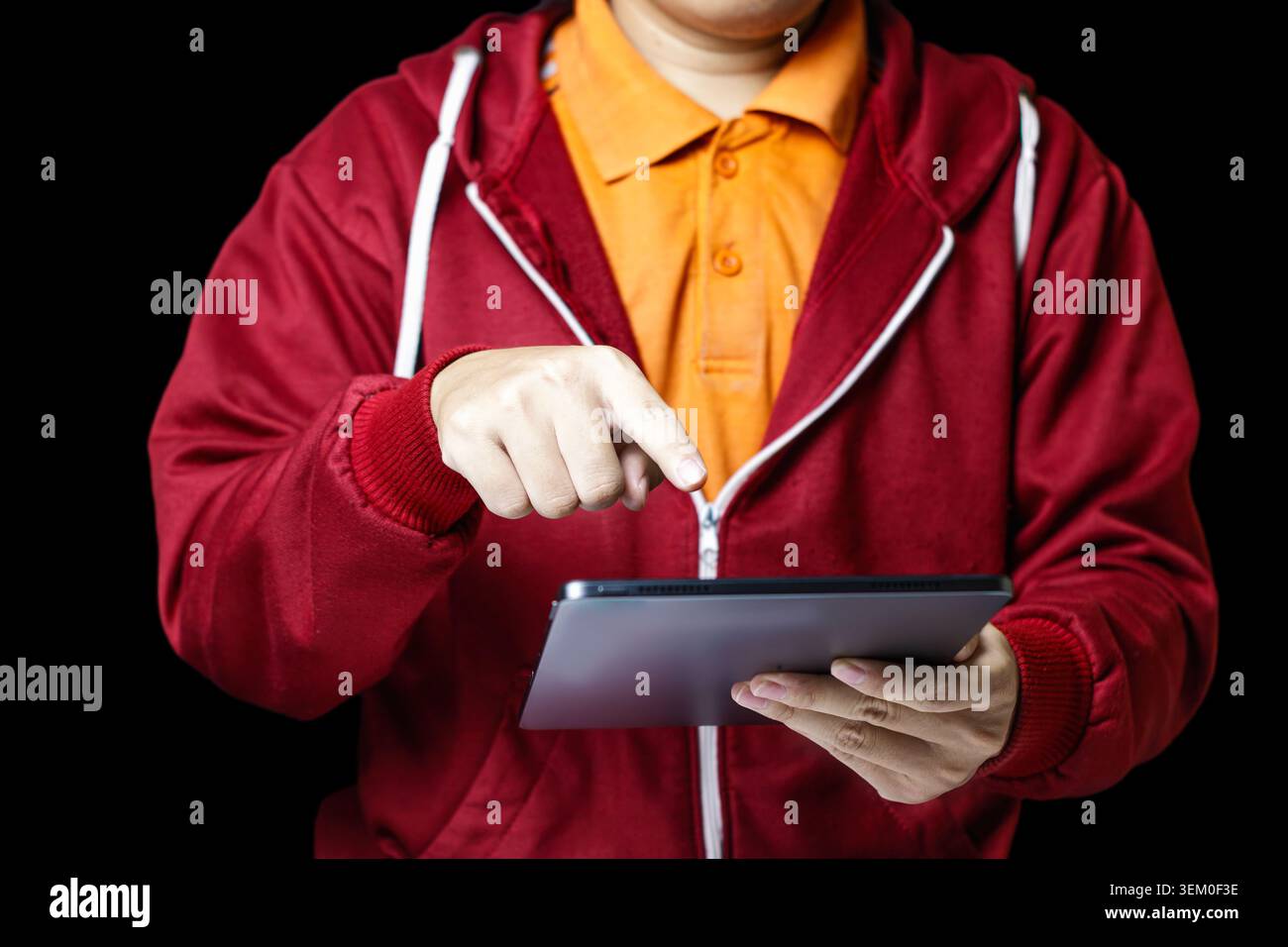 Homme en sweat à capuche rouge tient la tablette. Appuie avec le doigt sur l'écran avec l'intention mise au point. Chemise orange vif contraste le fond sombre. Une tenue élégante suggère un utilisateur moderne et doué de technologie. Idéal pour les affaires, l'éducation. Photo de haute qualité Banque D'Images