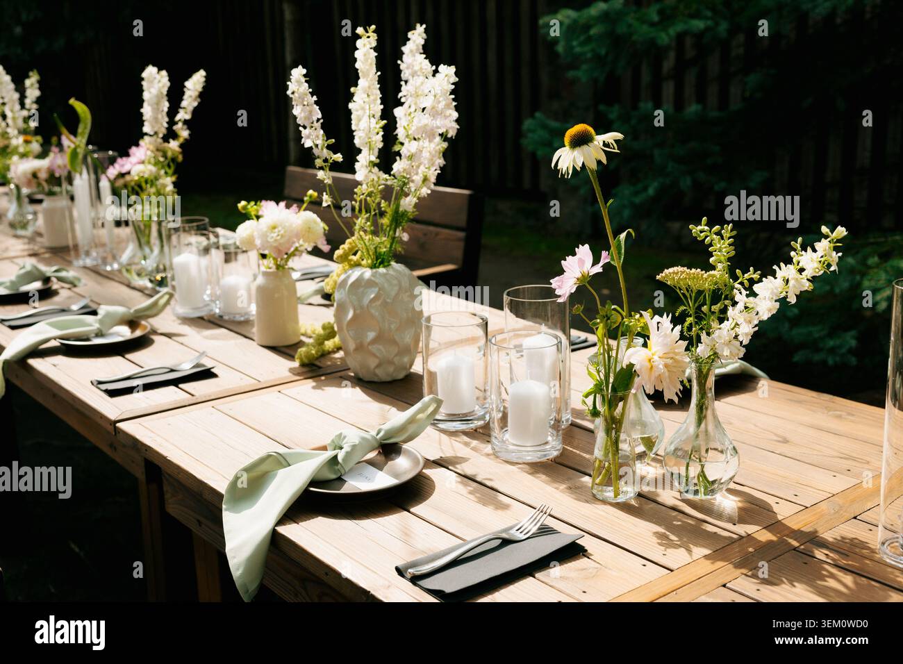 Table de ferme de mariage en plein air rustique avec vases à bourgeons de fleurs sauvages, bougies piliers, serviettes vert sauge et décors d'endroit noir dans la lumière du soleil de jardin tapissé. Banque D'Images