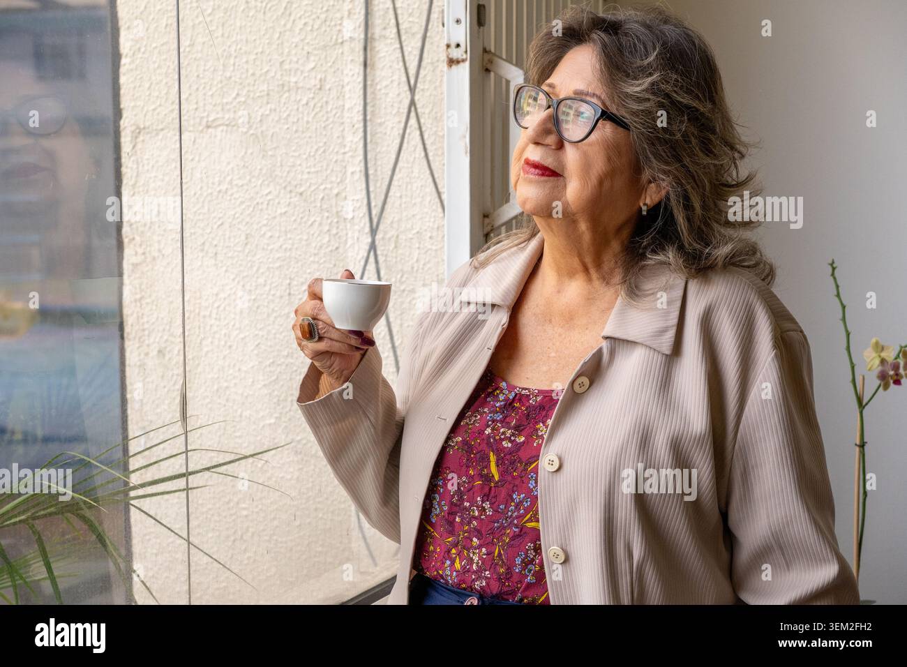 Femme senior regardant par la fenêtre, tenant une tasse de café, réfléchissant sur les pensées Banque D'Images