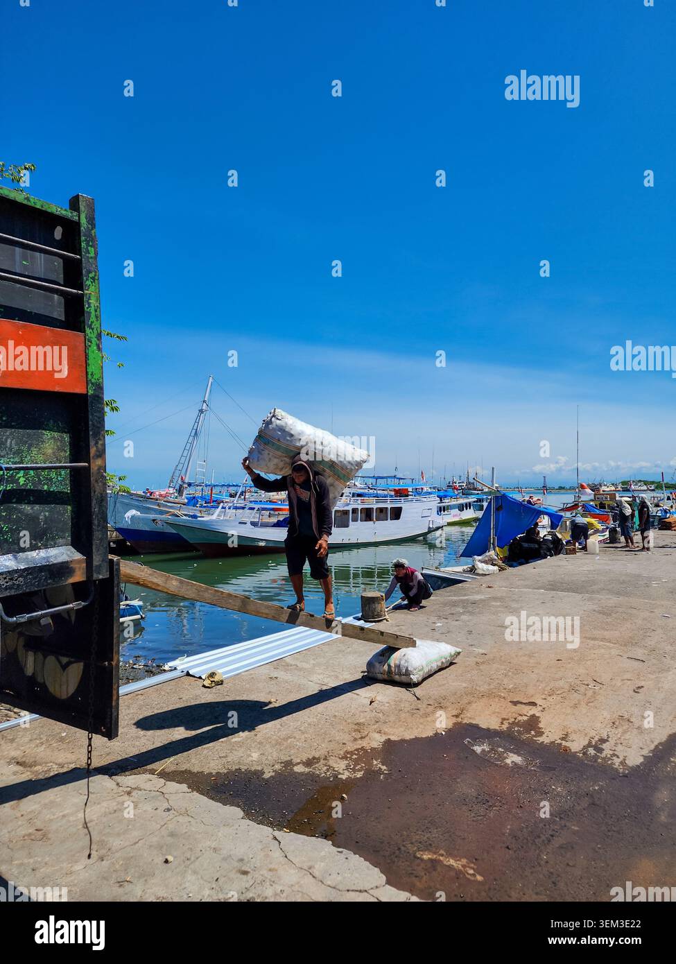 Makassar, Indonésie - 09 février 2024 : Ouvrier portant un sac sur une planche de bois au port de Paotere avec des bateaux traditionnels en arrière-plan. Banque D'Images