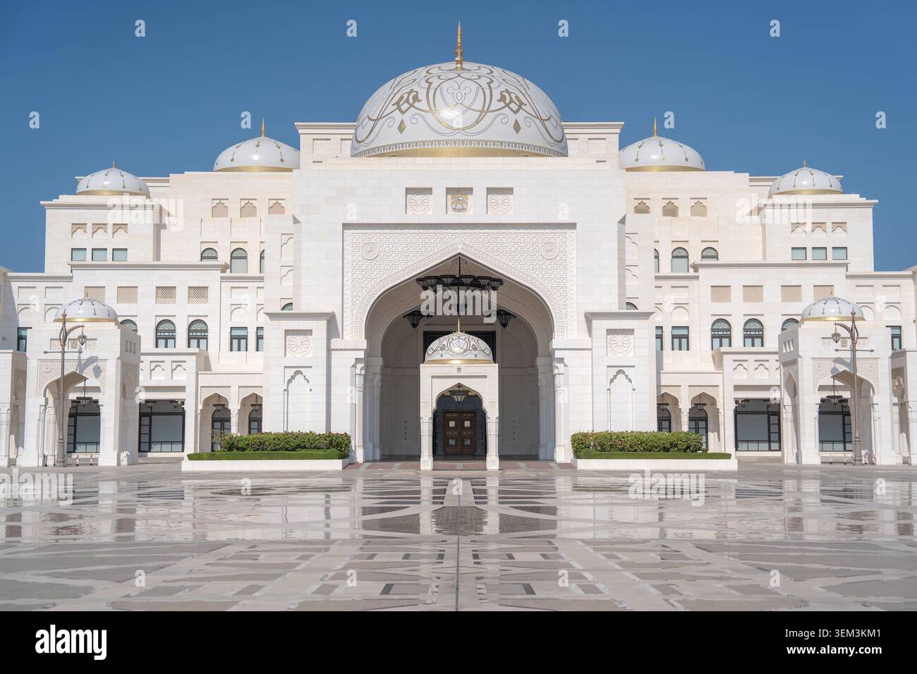 Bâtiment blanc semblable à un palais avec des dômes, des arches et une grande entrée, vu d'une cour carrelée brillante sous un ciel bleu clair. Banque D'Images