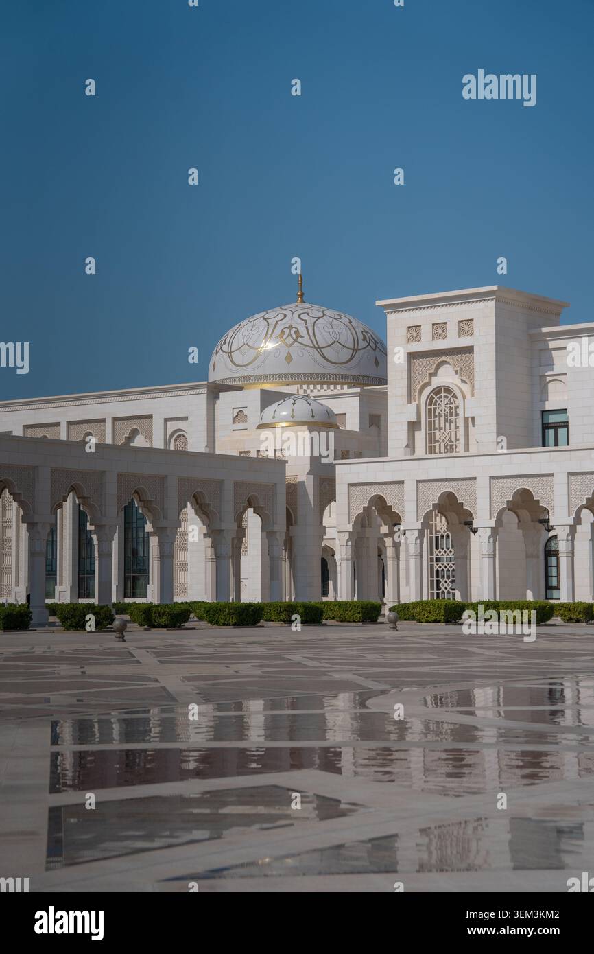 Mosquée ornée avec des arches blanches, un toit en dôme et une grande cour réfléchissant sur un sol humide sous un ciel bleu clair Banque D'Images