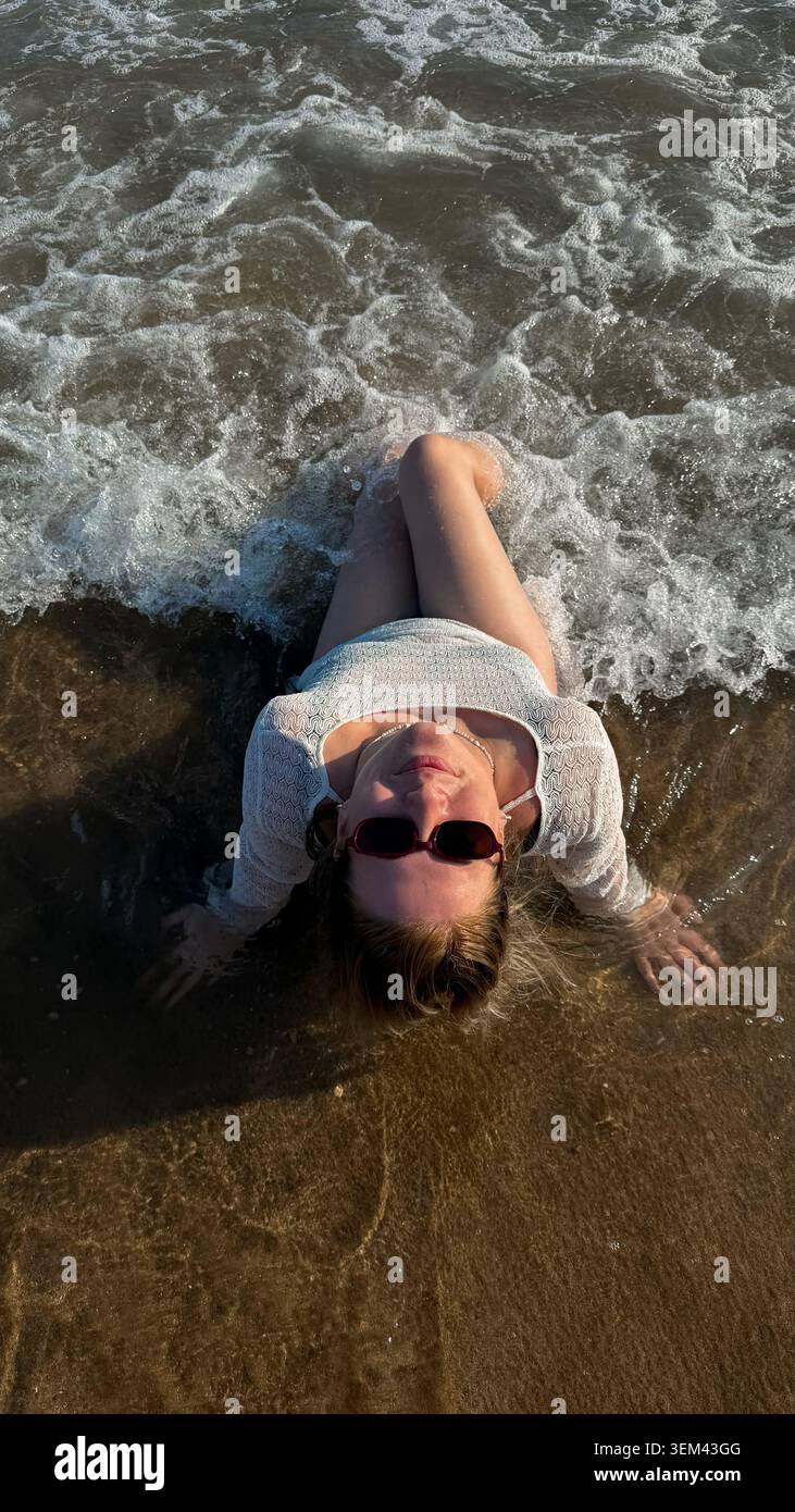 Jeune femme en lunettes de soleil couchée sur une plage de sable, partiellement submergée par les vagues de l'océan, profitant d'une journée ensoleillée avec des éclaboussures d'eau autour d'elle Banque D'Images