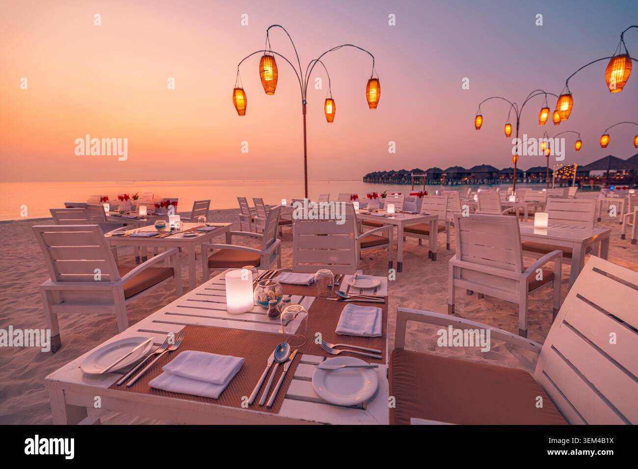 Restaurant en bord de mer avec des lanternes lumineuses au coucher du soleil, tables blanches dressées pour le dîner sur le sable, villas sur pilotis à l'horizon, dîner dans un resort tropical de luxe Banque D'Images