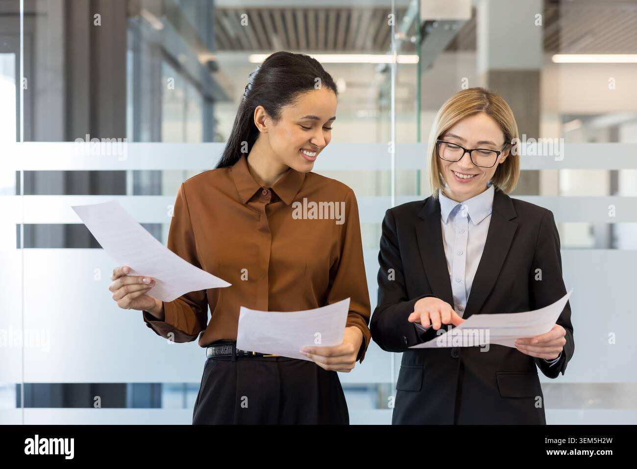 Deux femmes d'affaires souriantes dans un bureau moderne collaborent sur des documents et des graphiques, discutent de stratégie et analysent les données alors qu'elles planifient un projet et célèbrent ensemble les progrès Banque D'Images
