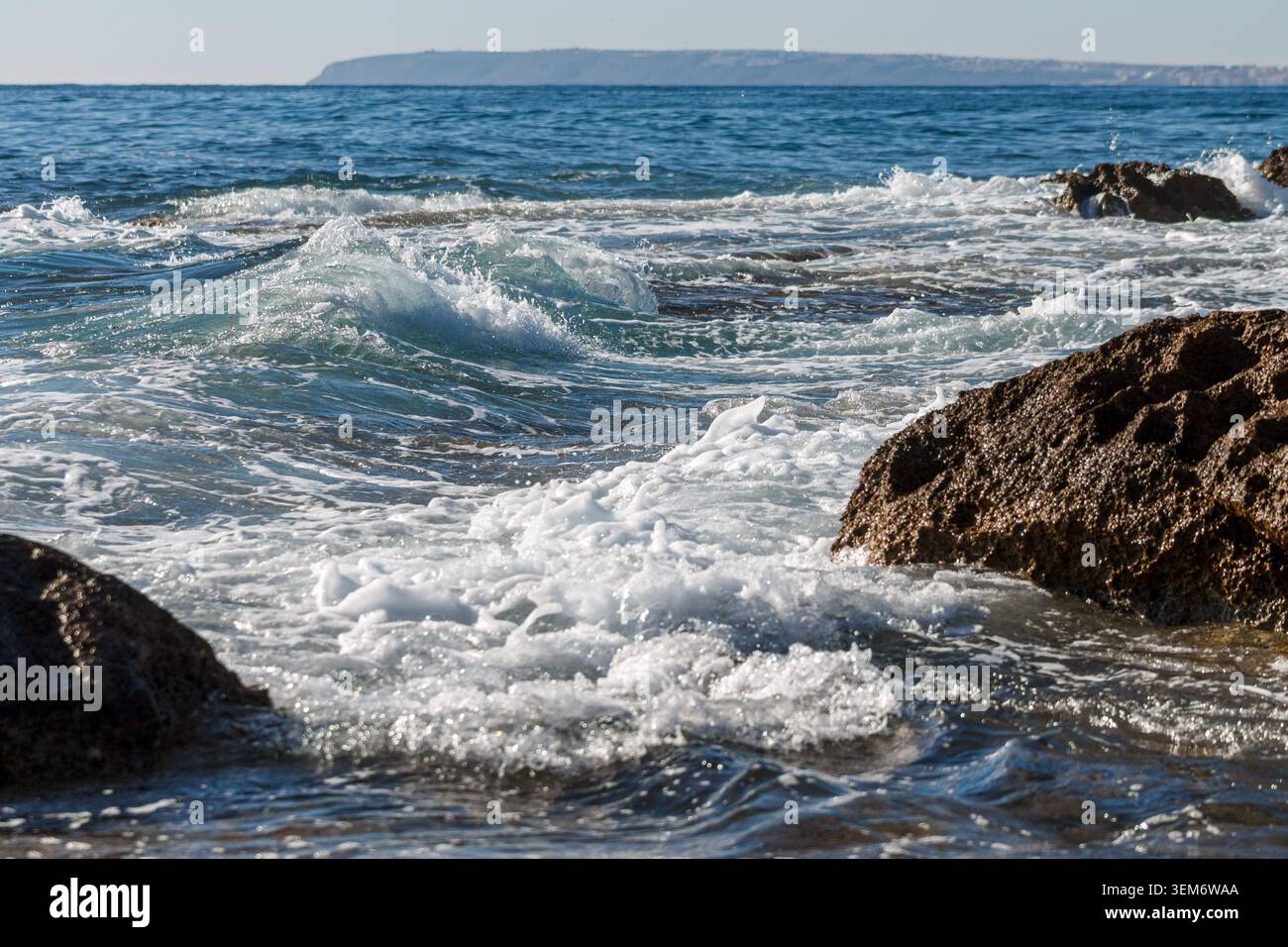 Plan de jour des vagues qui s'écrasent sur la côte rocheuse au premier plan, et la colline de Santa Pola en arrière-plan, sur le littoral espagnol de la costa blanca Banque D'Images