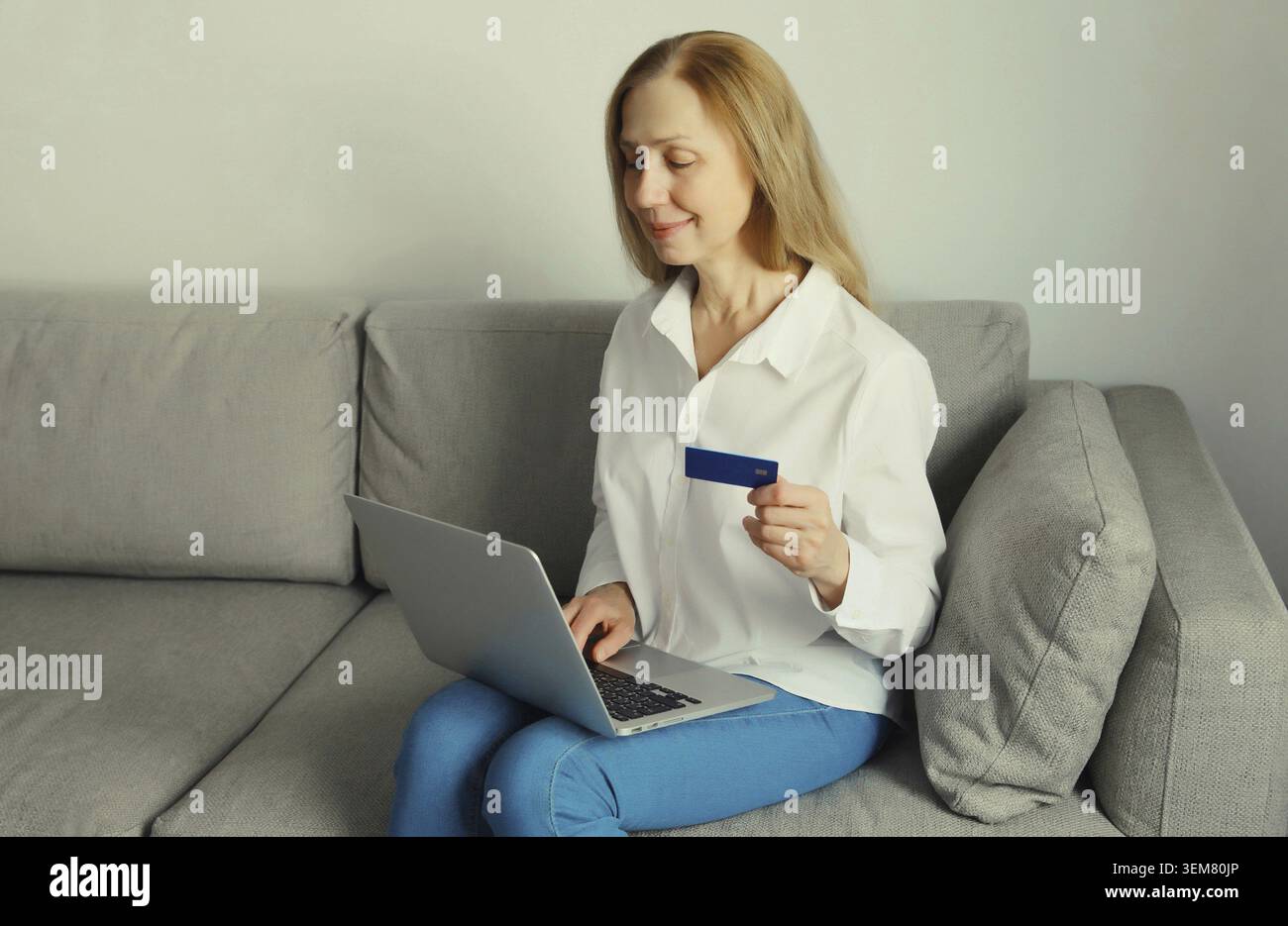 Heureuse femme souriante utilisateur tenant la carte bancaire de crédit en plastique, regardant l'ordinateur portable, magasinant en ligne ou commandant des marchandises via l'ordinateur assis sur le canapé en L. Banque D'Images