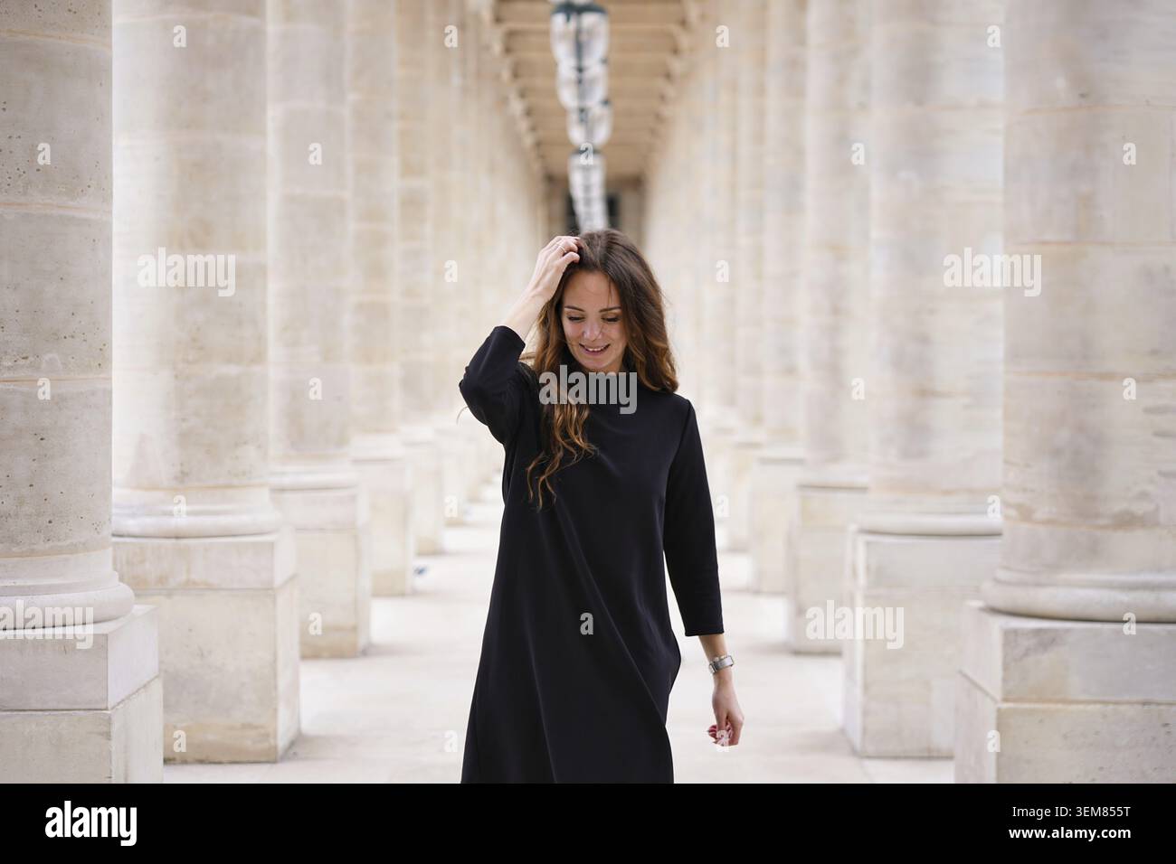 Jeune femme joyeuse en robe noire marchant en toute confiance à travers la colonnade architecturale classique au Palais Royal à Paris, France Banque D'Images