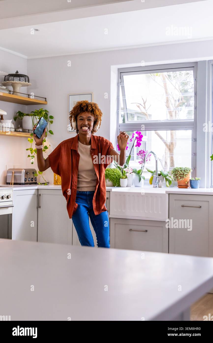 Femme afro-américaine dansant et tenant smartphone dans la cuisine à la maison lumineuse près de l'évier, orchidée rose Banque D'Images