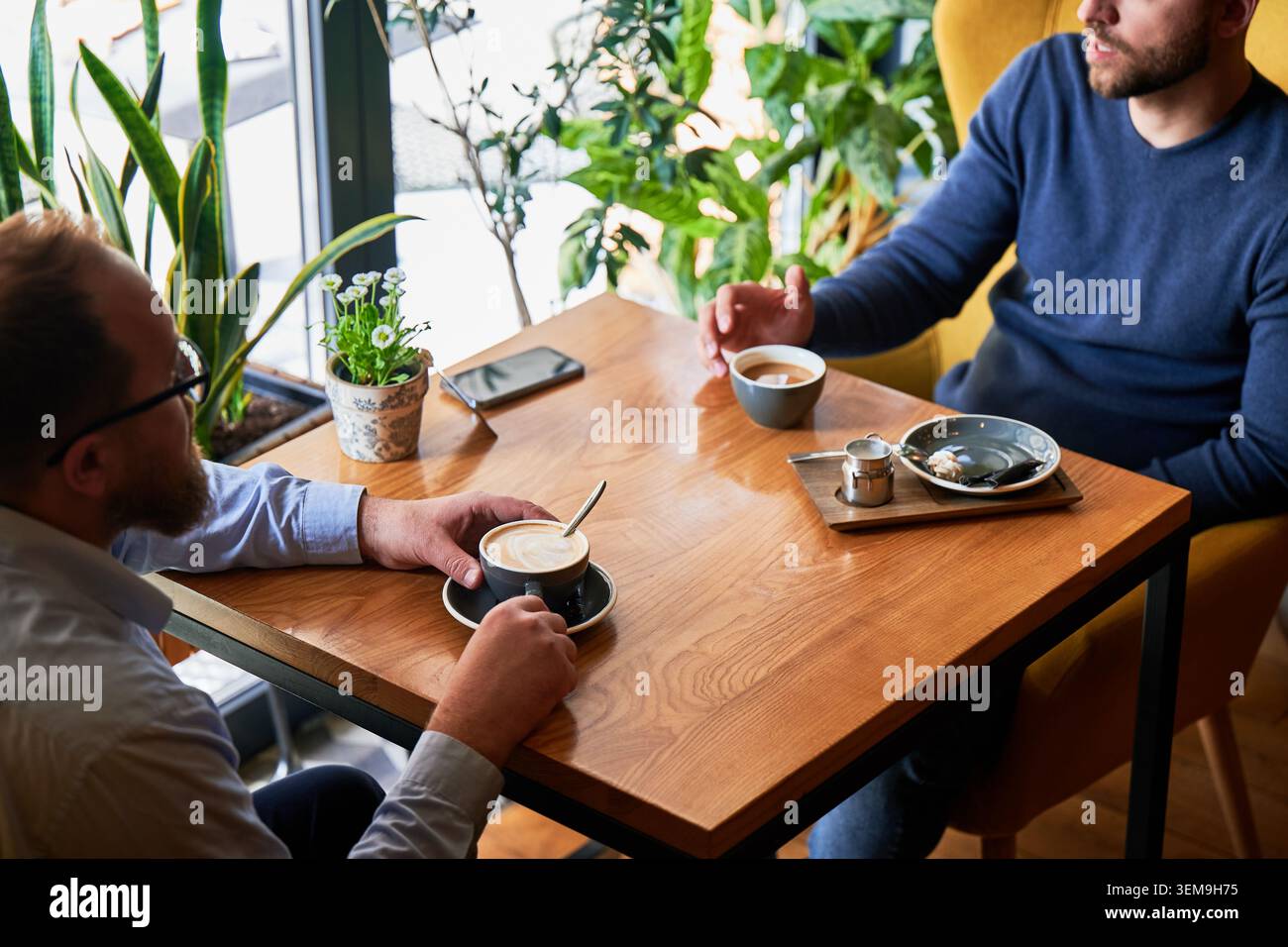 Deux hommes ayant une conversation de café à la table de café, réunion informelle ou chat amical, atmosphère détendue avec des boissons, smartphone sur la table et plantes vertes dans un intérieur lumineux Banque D'Images