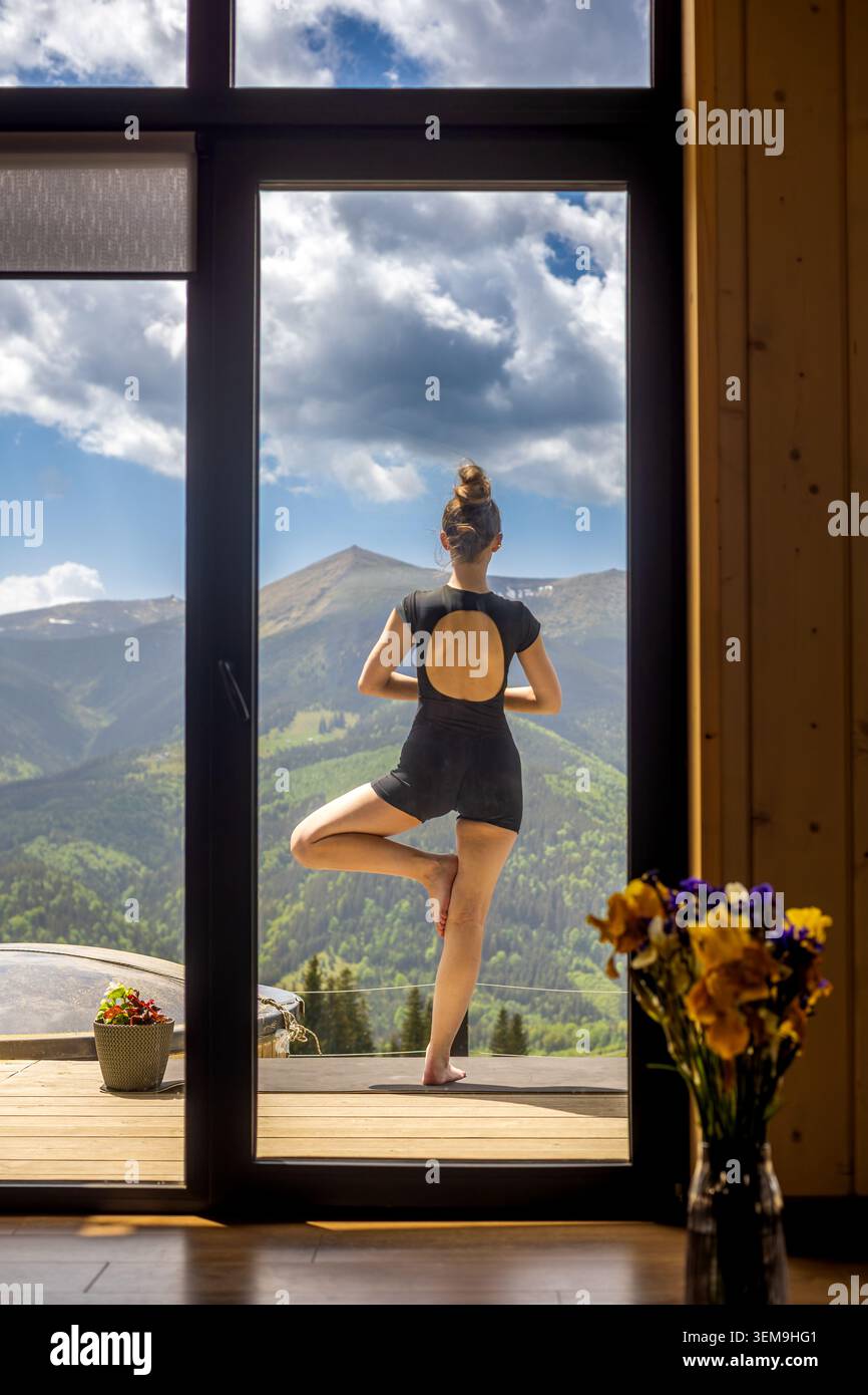 Femme pratiquant la pose d'arbre avec vue sur la montagne. Femme en Activewear noir effectue la pose d'arbre sur une terrasse, encadrée par une fenêtre de cabine avec pittoresque alpin Banque D'Images