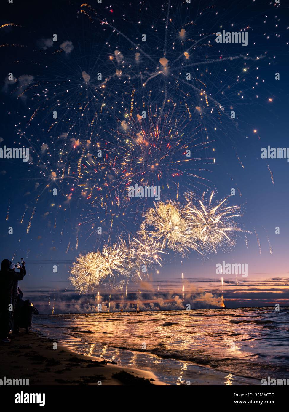 Spectaculaire feu d'artifice doré illuminant le ciel nocturne et se reflétant sur les vagues de la mer lors d'un événement estival côtier en Europe. Banque D'Images