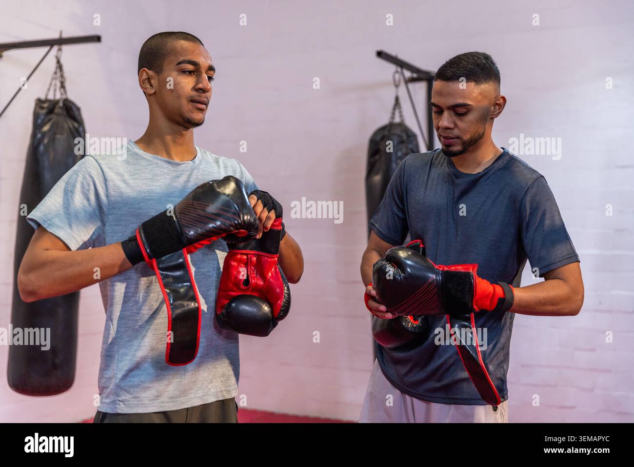 Deux hommes ajustant des gants rouges-noirs et des enveloppements dans des vêtements de sport près de sacs suspendus dans la salle de boxe Banque D'Images