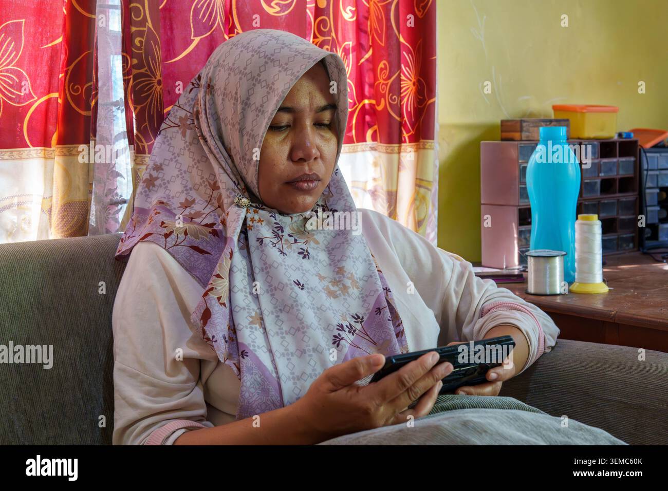 Une femme musulmane portant un hijab interagit avec l'écran de son smartphone lorsqu'elle est assise sur un canapé à l'intérieur à Mataram, Lombok, West Nusa Tenggara. Banque D'Images