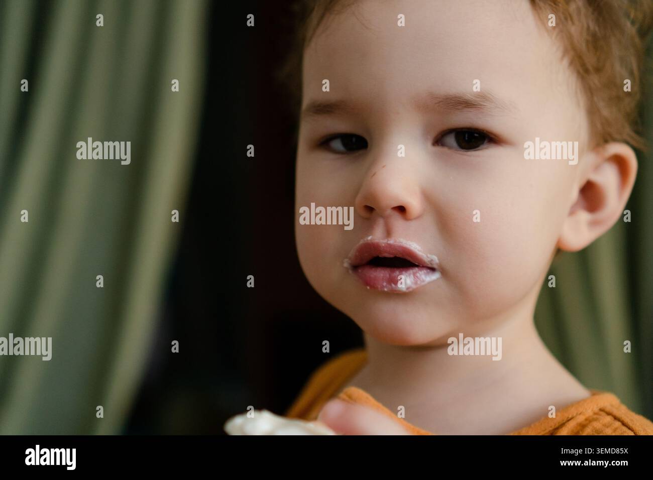 Adorable garçon avec de la nourriture sur la lèvre regarde la caméra en gros plan portrait. Tout-petit ludique montre le désordre innocent de l'enfance tout en mangeant à la maison. Doux moment attachant avec une expression naturelle non posée à l'intérieur Banque D'Images