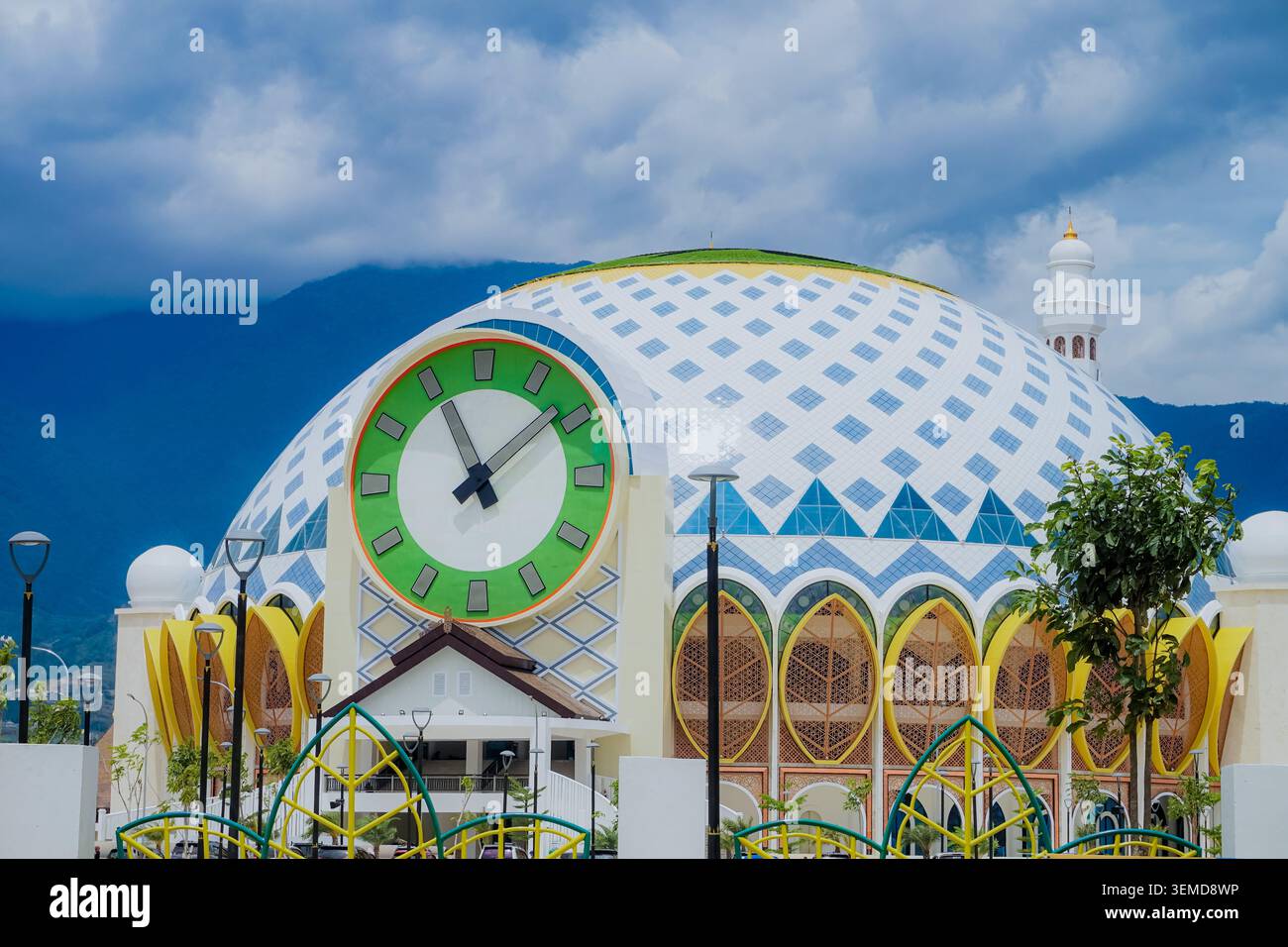 palu, Indonésie - 10 mars 2026 : façade avant de la Grande Mosquée Baitul Khairaat présentant une horloge analogique massive encastrée dans un dôme à motif de losanges, de grandes fenêtres jaunes arquées et un fond montagneux par temps nuageux. Banque D'Images