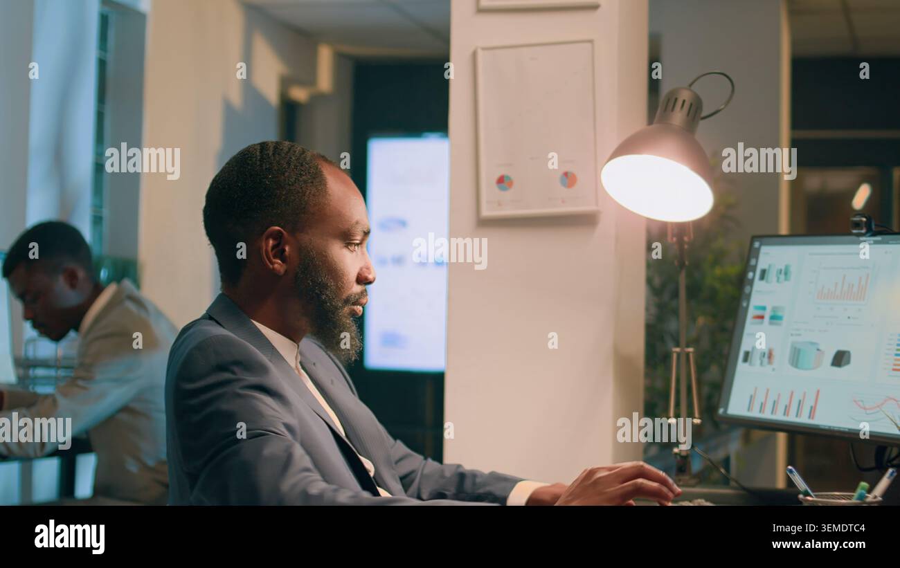 Hommes d'affaires afro-américains assis à leur bureau, regardant sur des graphiques financiers, imputant des données sur ordinateur pendant le quart de nuit. Collègues résolvant des tâches de projet d'entreprise du jour au lendemain avant la date limite Banque D'Images