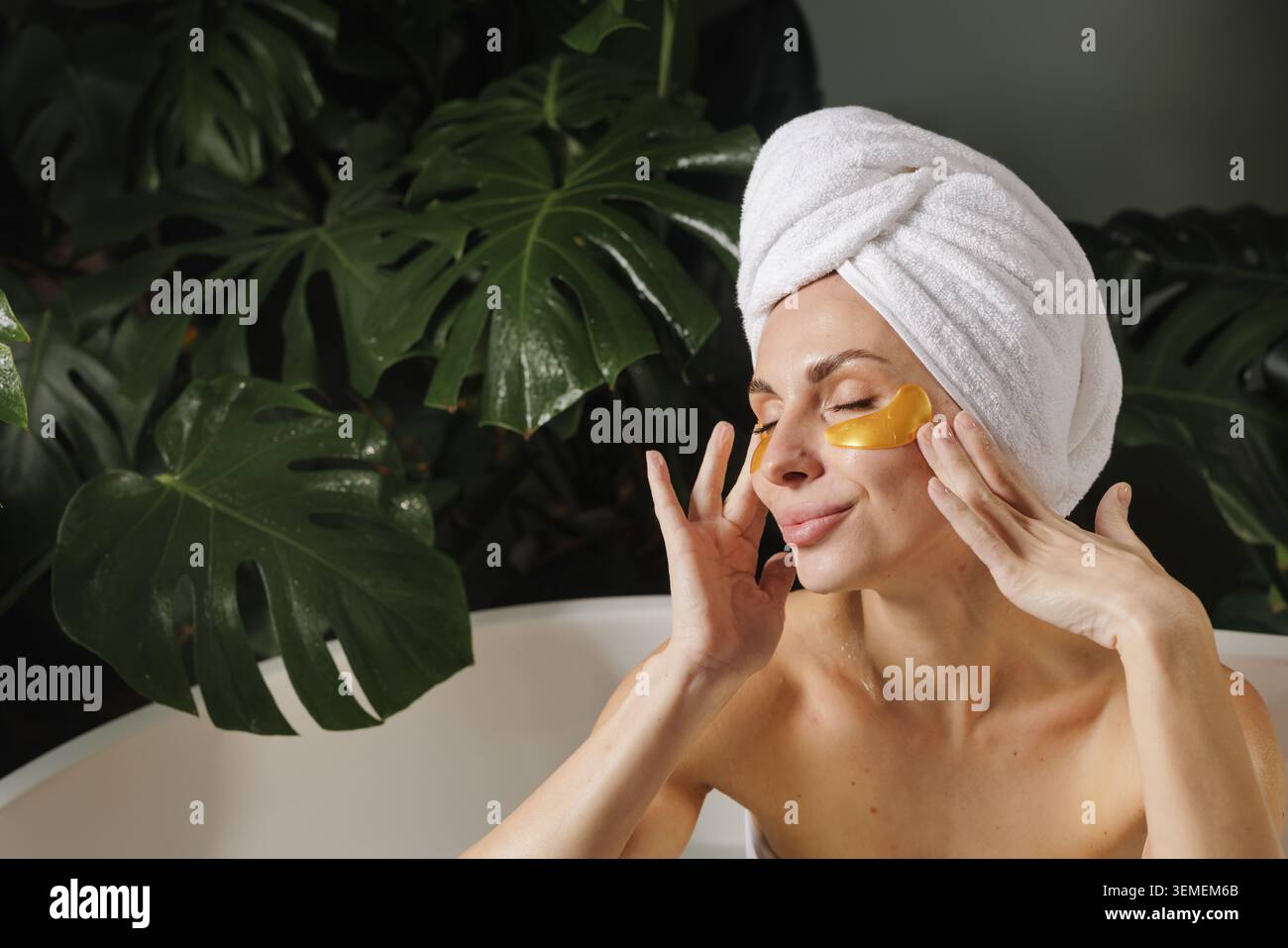 Une femme se détend dans une salle de bain sereine, en appliquant des patchs oculaires dorés tout en profitant d'un moment d'auto-soin entouré de plantes vertes luxuriantes. Banque D'Images