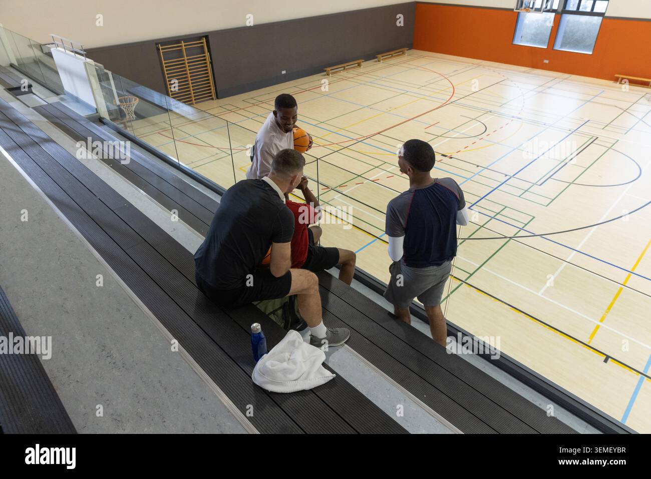Divers coéquipiers masculins dans l'équipement de sport parlant tout en étant assis sur des gradins à la salle de gym de l'école avec balle Banque D'Images