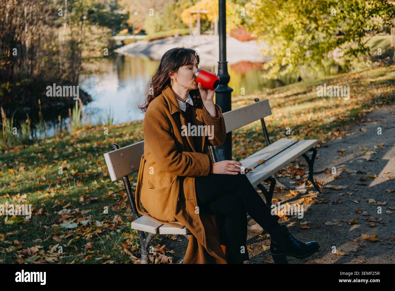 Femme est assise sur le banc du parc buvant dans une tasse réutilisable près de l'eau pendant l'automne. Routine automnale lente, escapade nature détox numérique, automne confortable Banque D'Images