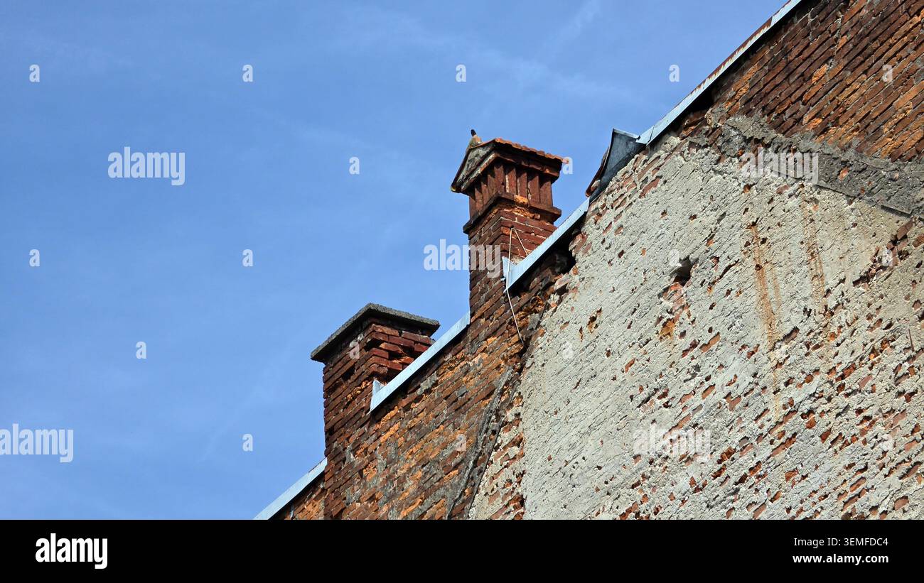 Deux cheminées en briques altérées s'étendent au-dessus de la ligne de toit d'un ancien bâtiment avec du plâtre en ruine et de la maçonnerie exposée sous un ciel bleu clair Banque D'Images