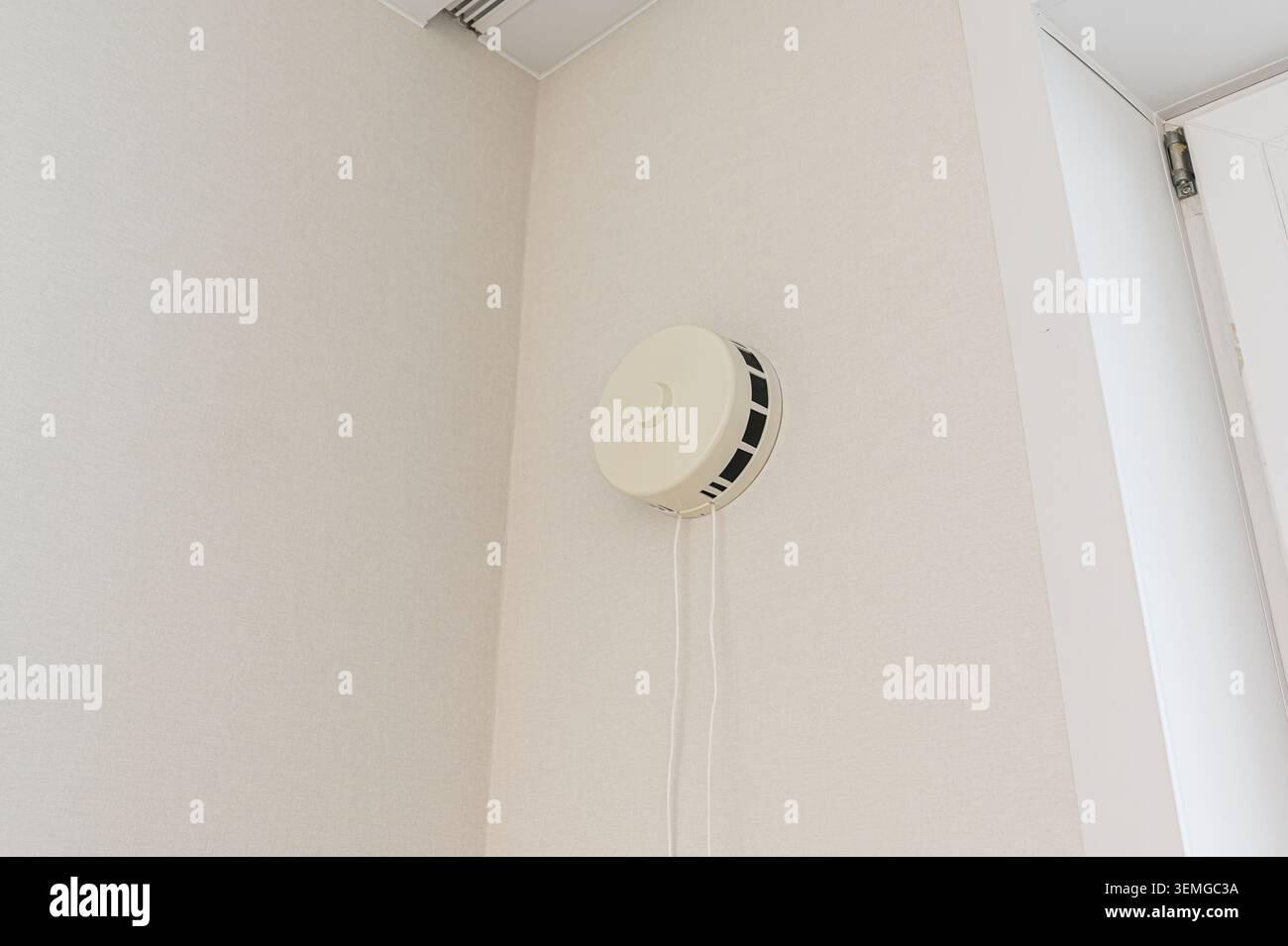 le détecteur de fumée circulaire blanc est monté sur un mur blanc dans une pièce. Les cordons pendent de l'appareil. Une partie d'une porte est vue. Banque D'Images