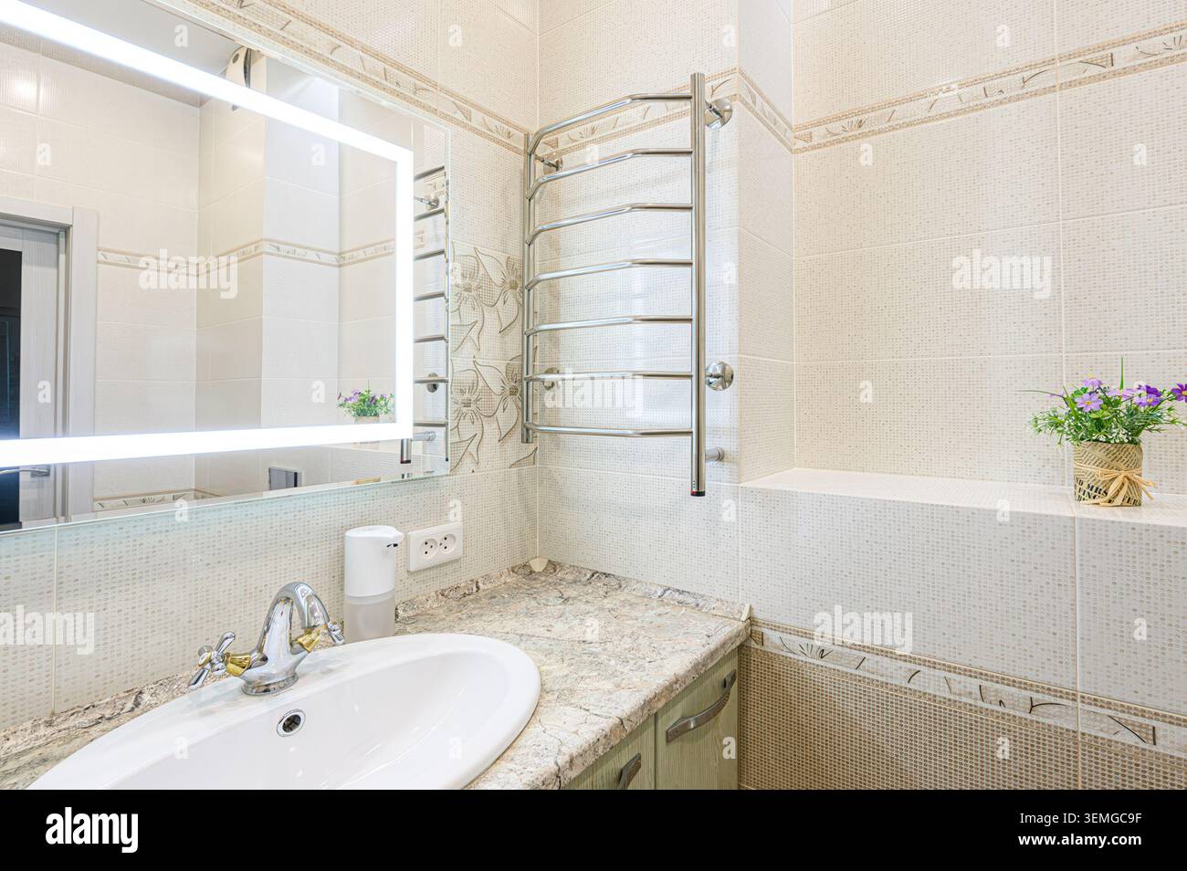 salle de bains bien éclairée avec carrelage beige, miroir lumineux moderne, lavabo avec comptoir en granit et sèche-serviettes. Une plante décorative en pot ajoute Banque D'Images