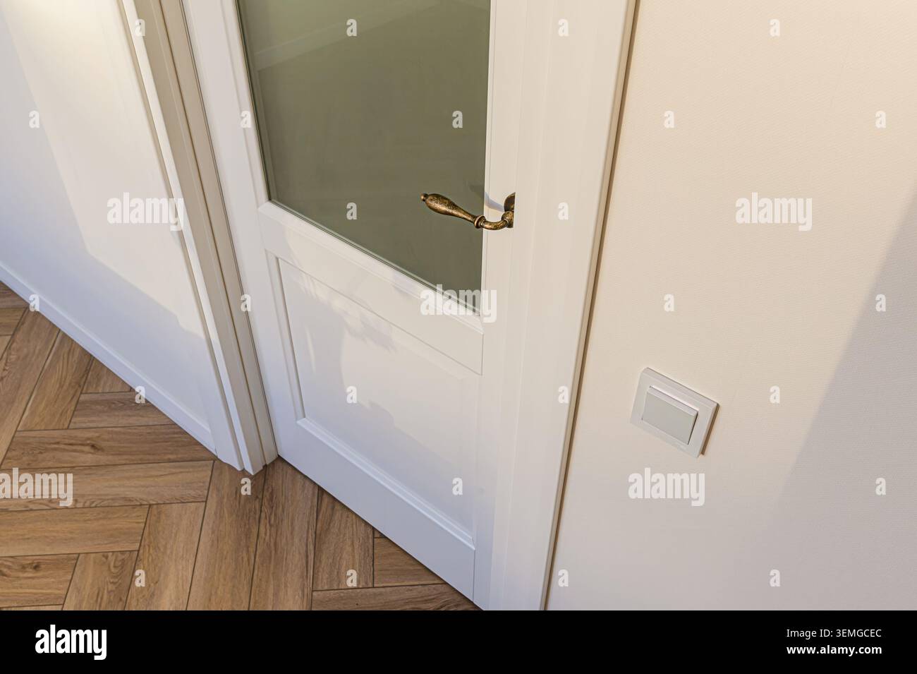 gros plan d'une porte intérieure blanche avec panneau de verre, poignée en bronze et interrupteur d'éclairage. Parquet aux tons chauds Banque D'Images
