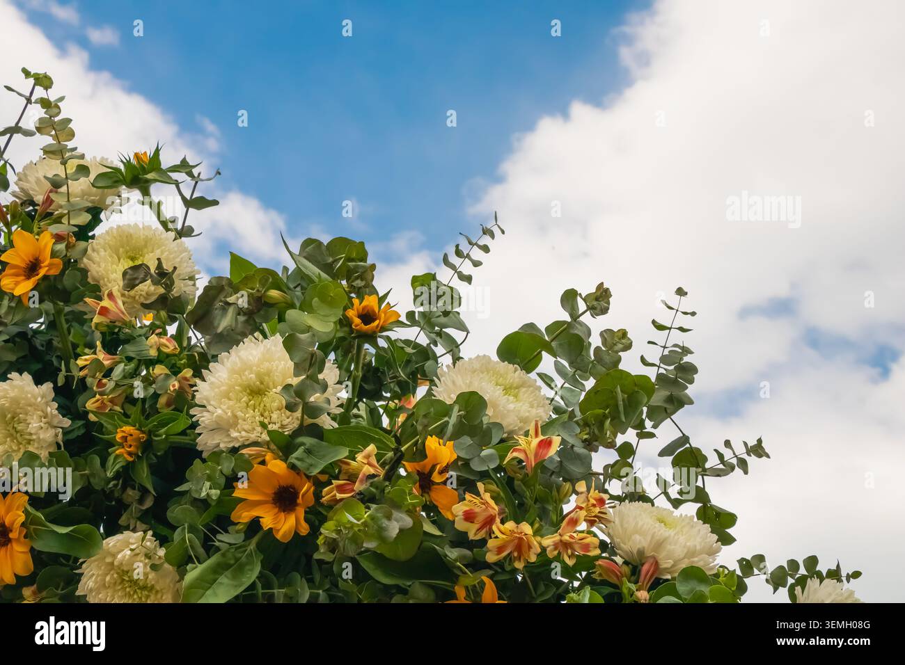 Arrangement floral décoratif avec chrysanthèmes blancs, fleurs jaunes et feuillage d'eucalyptus sous un ciel nuageux lumineux, idéal pour l'événement et Banque D'Images