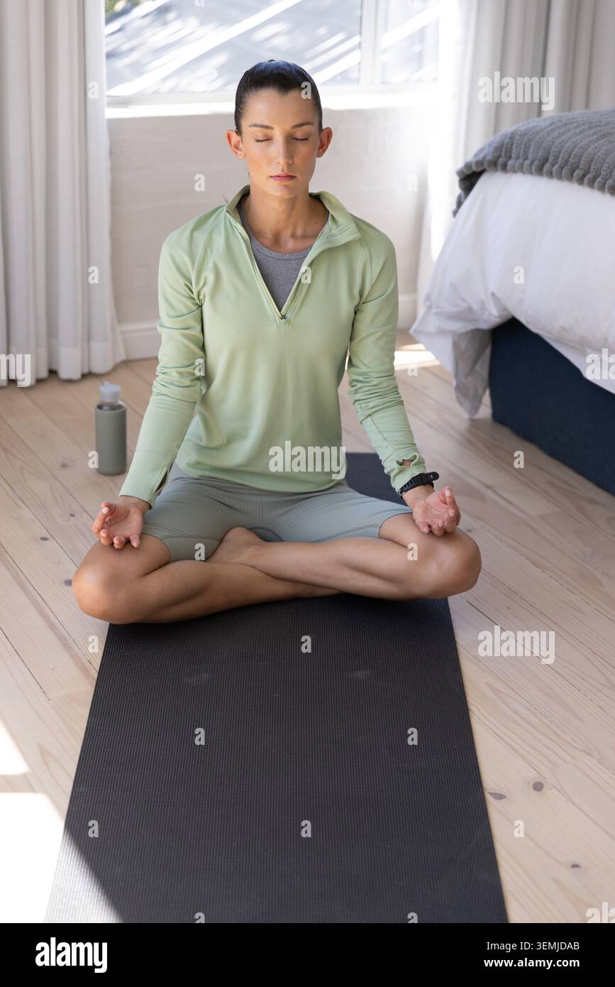Femme des années 30 méditant les jambes croisées sur un tapis de yoga noir dans la chambre à coucher par la fenêtre, portant un pull vert Banque D'Images